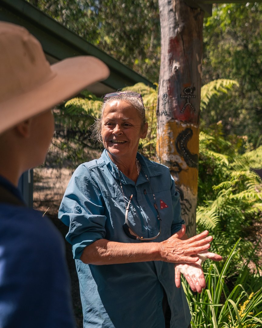 A woman with gray hair smiling and wearing a blue shirt, talking outdoors with another person partially visible in a tan hat, near a wooden pole with indigenous art, surrounded by greenery.