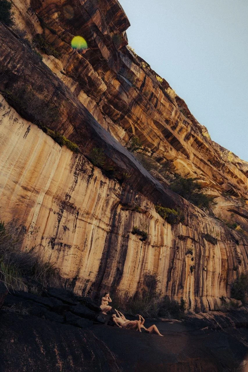Three people sunbathing on a rocky surface at the base of a tall, layered cliff with a dry landscape and sparse vegetation.
