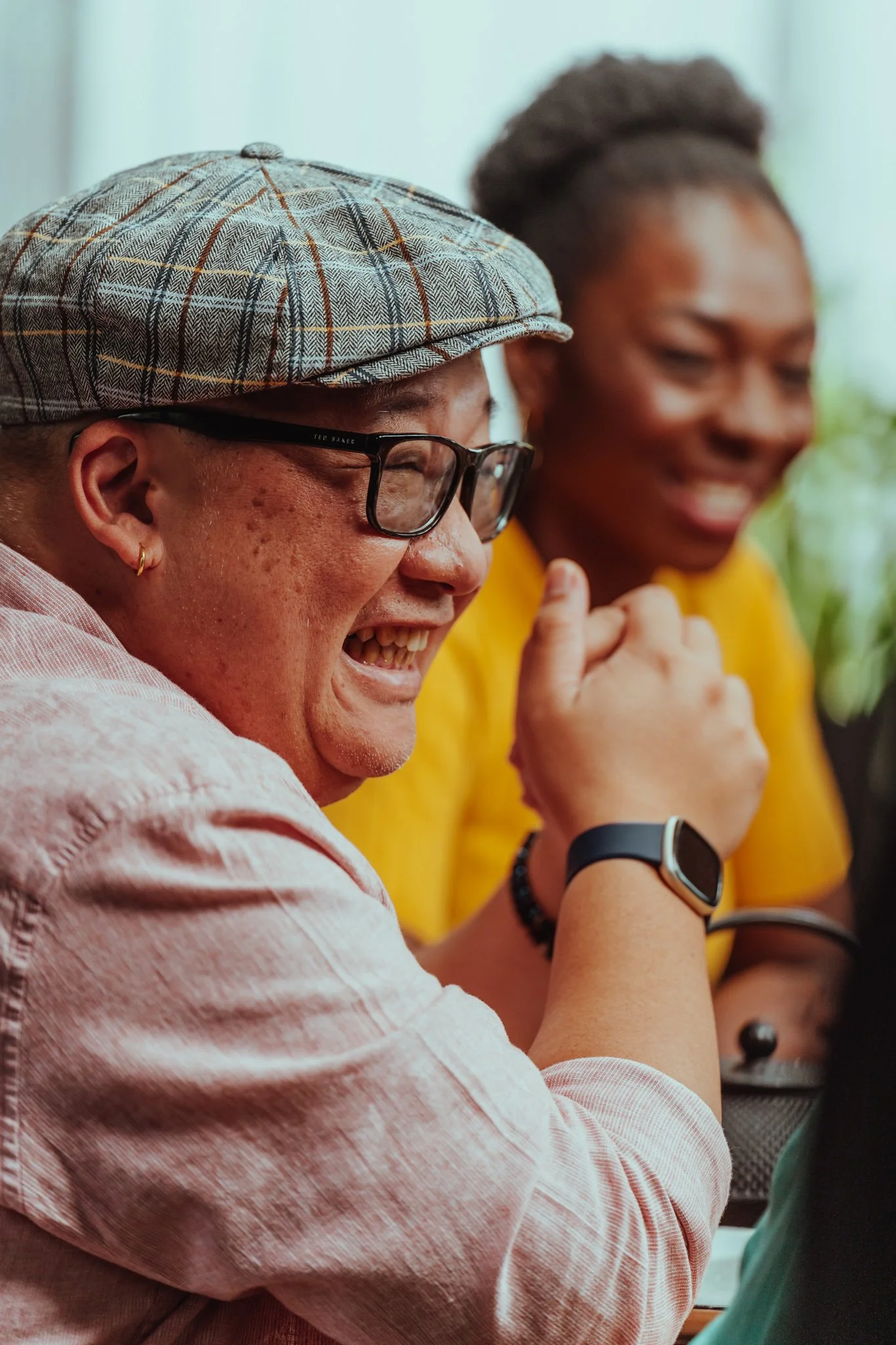 A man wearing glasses, a plaid cap, and a pink shirt, smiling and laughing, sitting next to a woman in a yellow shirt, also smiling, in a casual indoor setting.