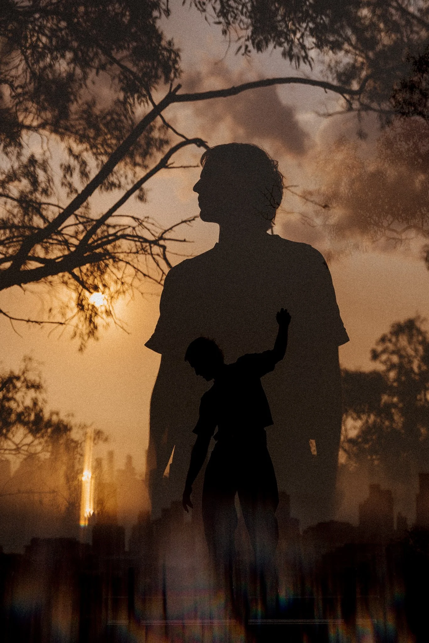 Silhouettes of two people, one larger and one smaller, against a sunset with trees and a cityscape in the background. The larger appears to be an adult, and the smaller a child, with the child raising one arm.