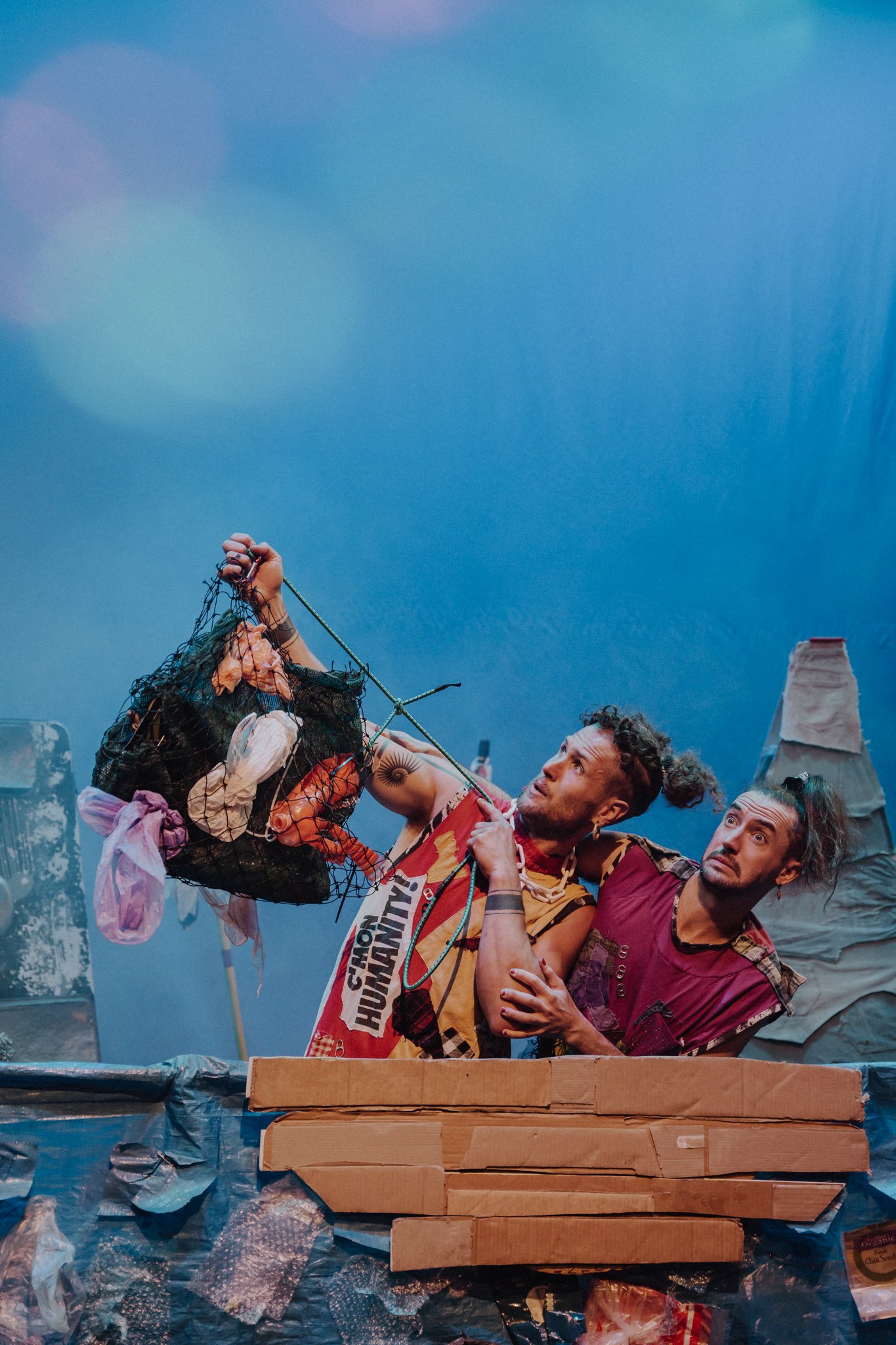 Two men with styled hair and expressive faces look at a bag filled with trash, amid a set resembling a junkyard with cardboard and paper structures, under a blue sky backdrop.