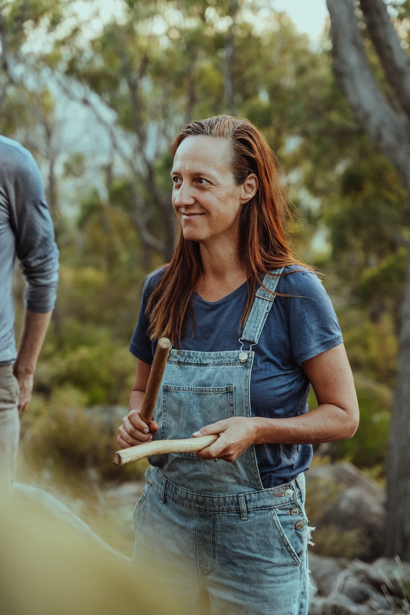 A woman with long red hair wearing a blue t-shirt and denim overalls holding a hatchet and small axe outdoors in a wooded area.