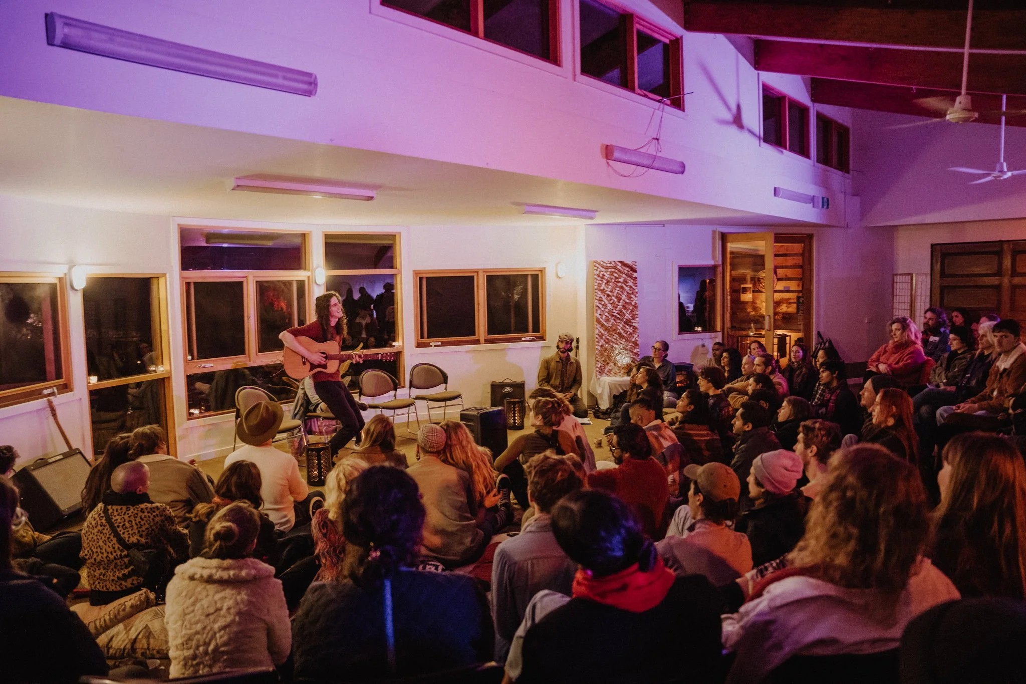 A woman playing an acoustic guitar and singing on a small stage in front of a seated audience in an indoor venue with wooden walls and ceiling fans, illuminated by warm lighting.