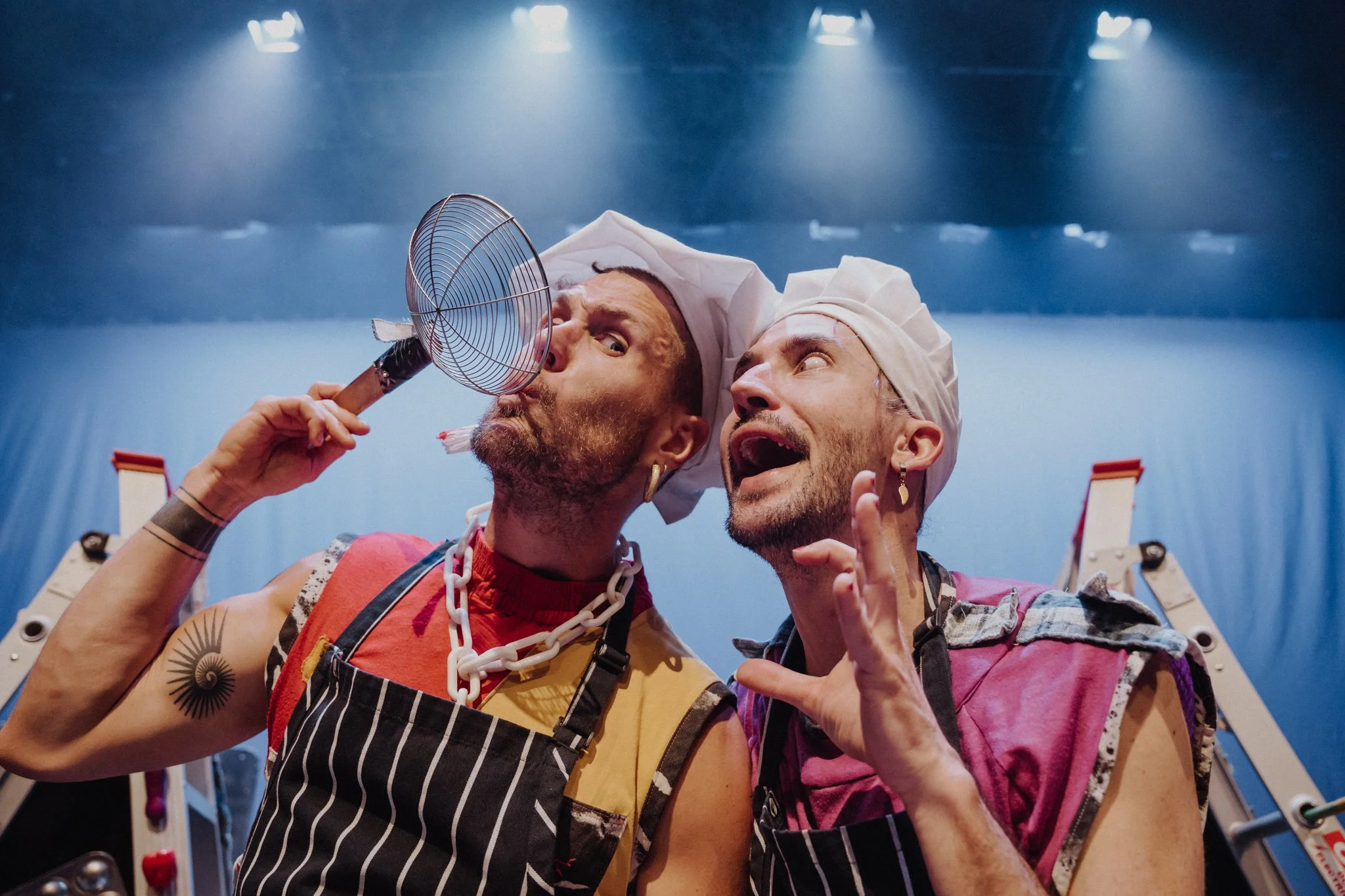 Two men wearing chef hats and aprons posing playfully on a stage, one pretending to blow a fan while the other gestures animatedly, with stage lighting overhead.