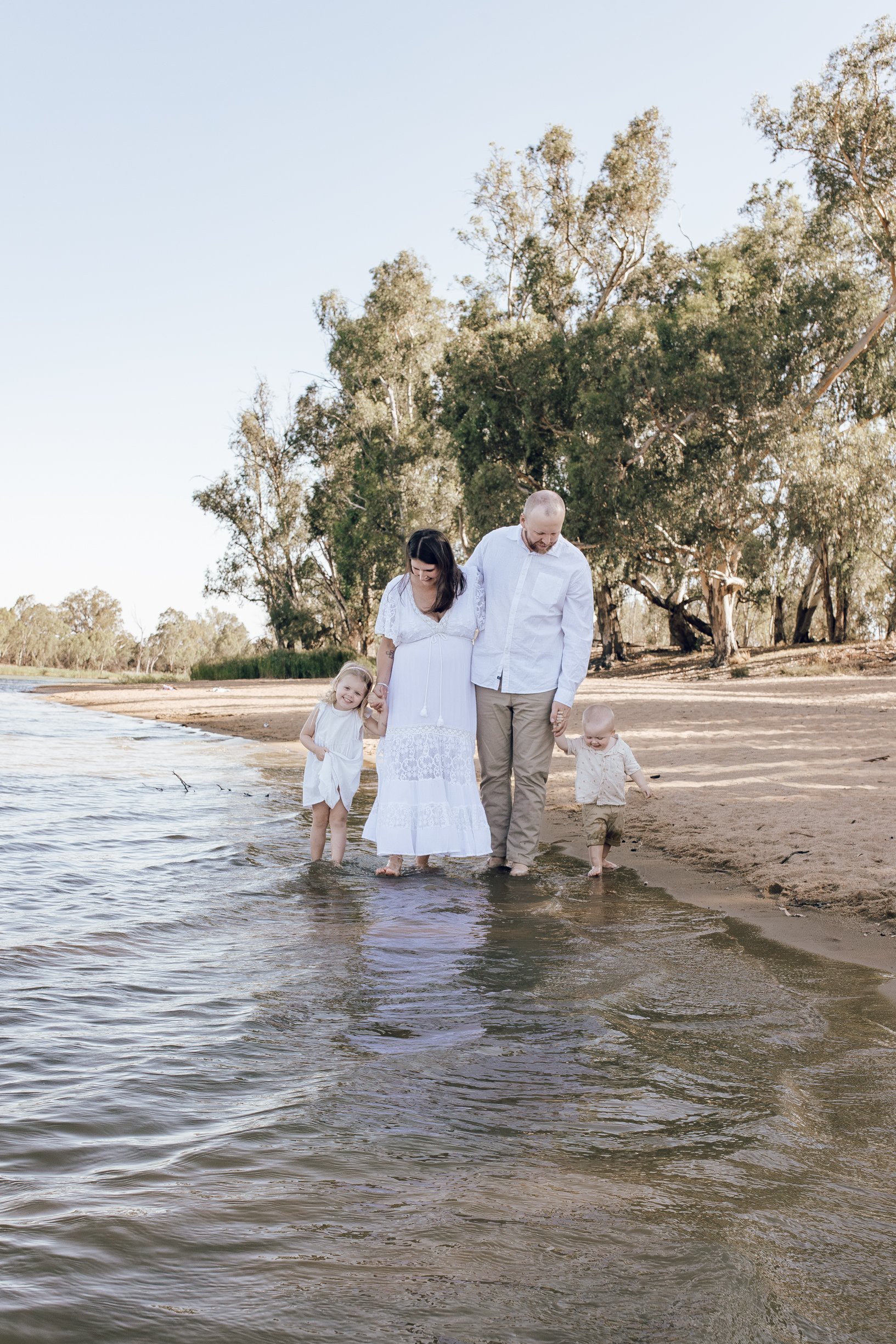 A family of four walking barefoot along the shoreline of a beach, holding hands, with trees in the background and the sky above.
