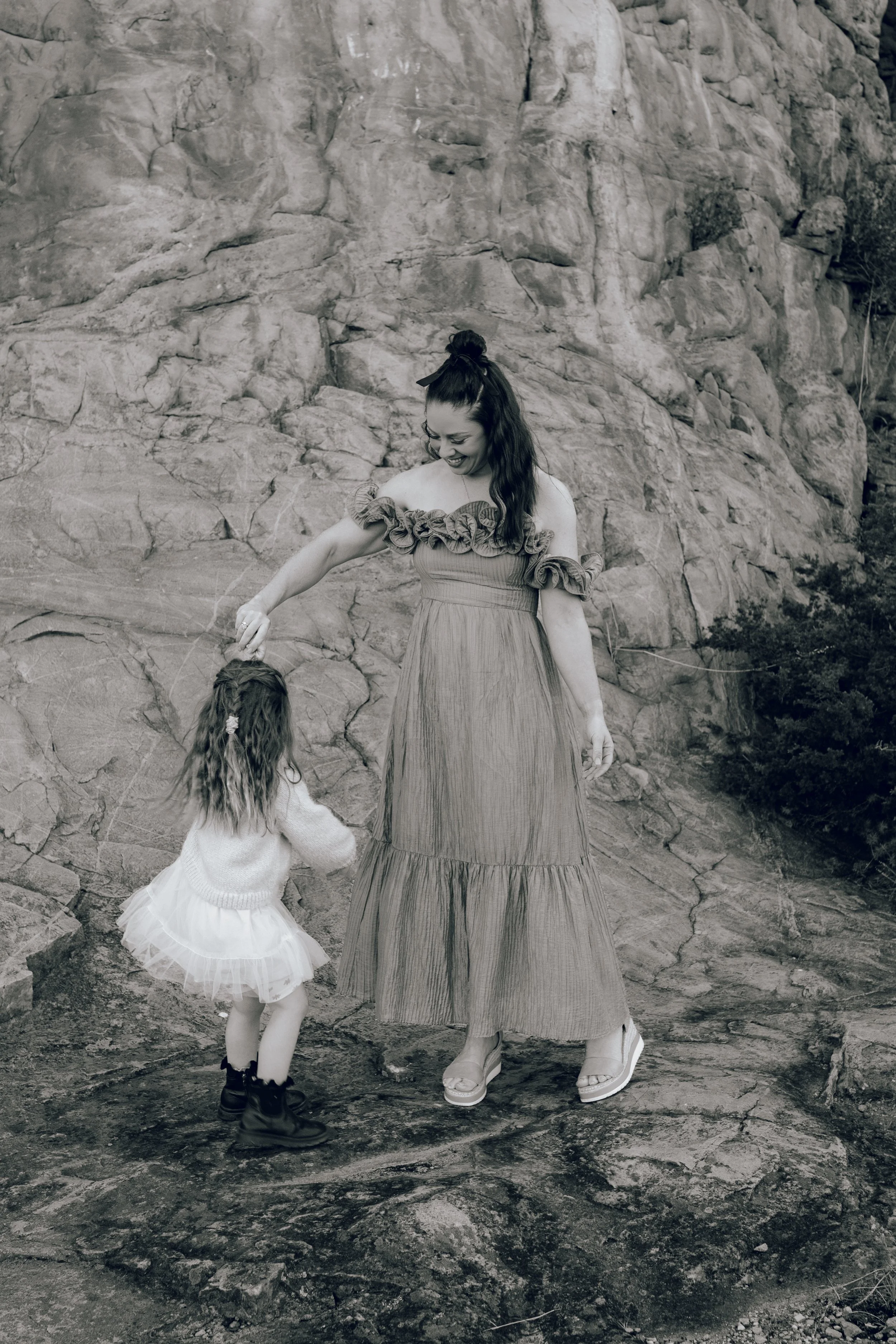 A woman and a young girl holding hands on a rocky outdoor surface with a large rock formation behind them.