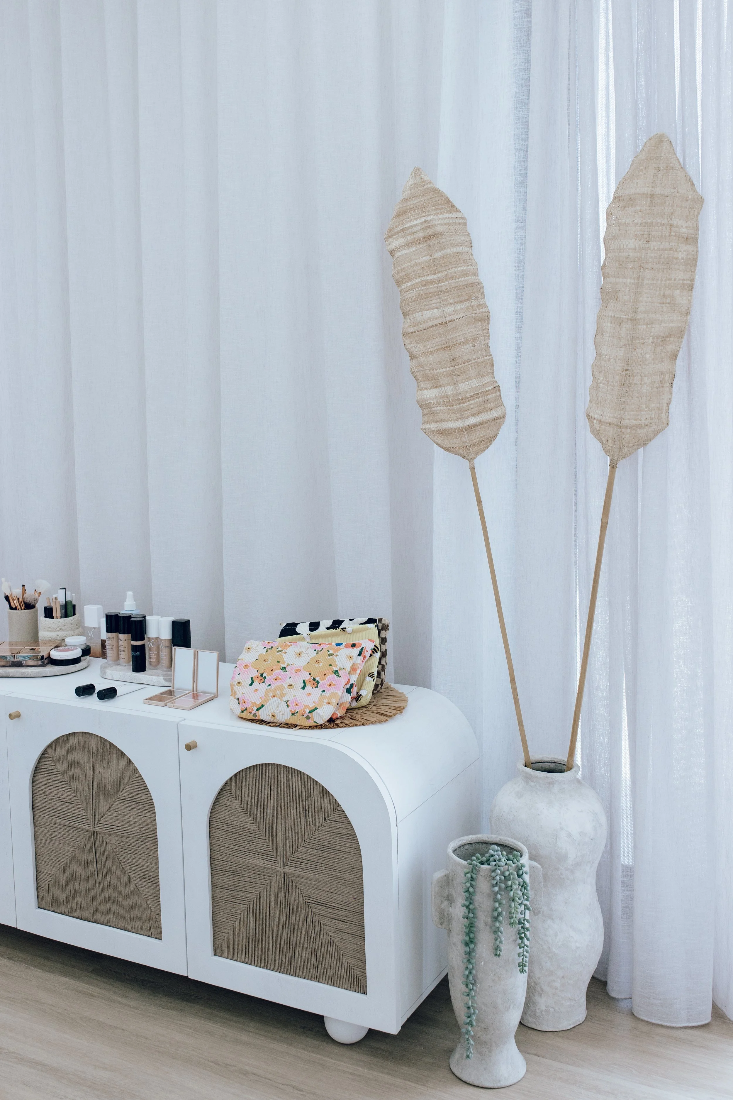 A white cabinet with rattan details, topped with makeup and skincare products, and decorative items, next to two large beige feathered plants in white vases, with a sheer curtain background.