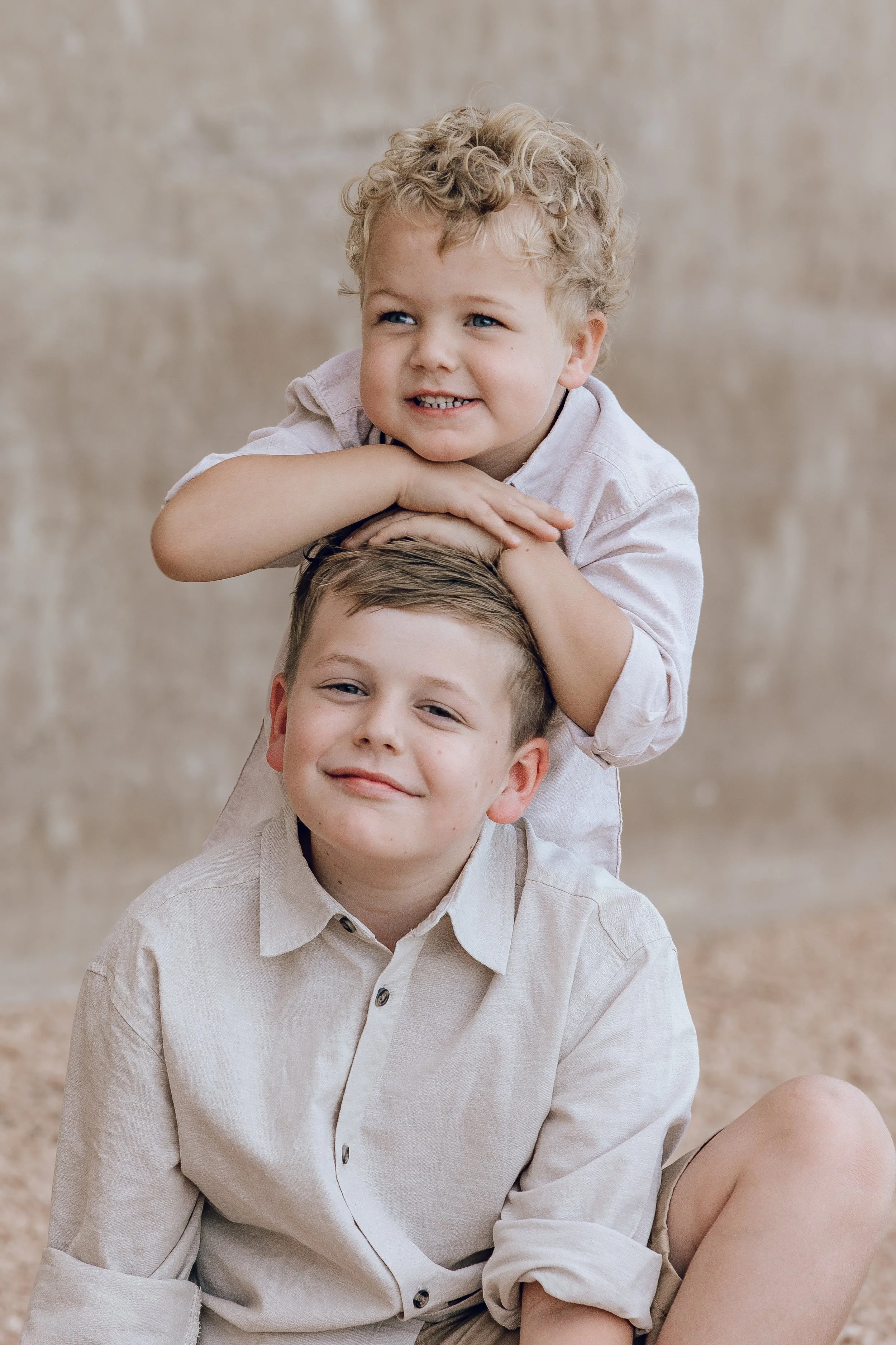 Two boys, one younger with curly hair resting his chin on folded hands atop the head of an older boy with straight hair, both smiling softly against a neutral background.