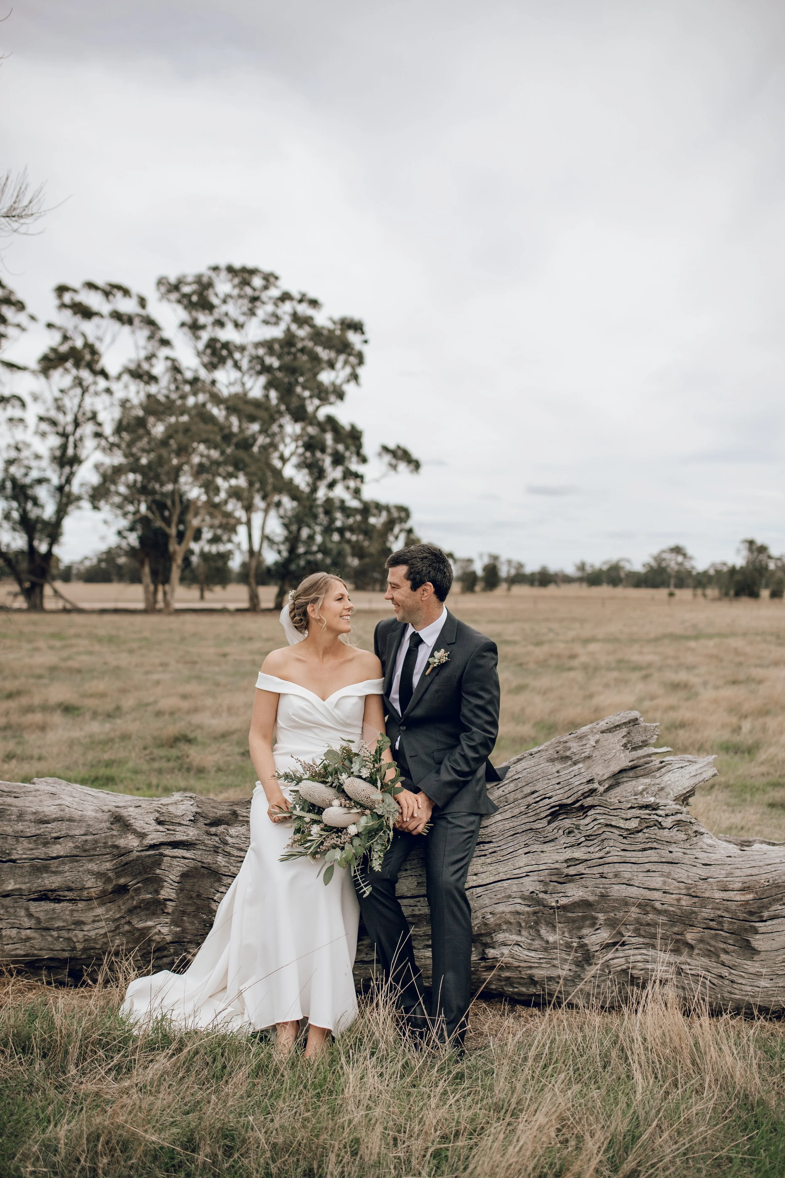 Bride and groom sitting on a fallen log in a field, smiling at each other with trees in the background.