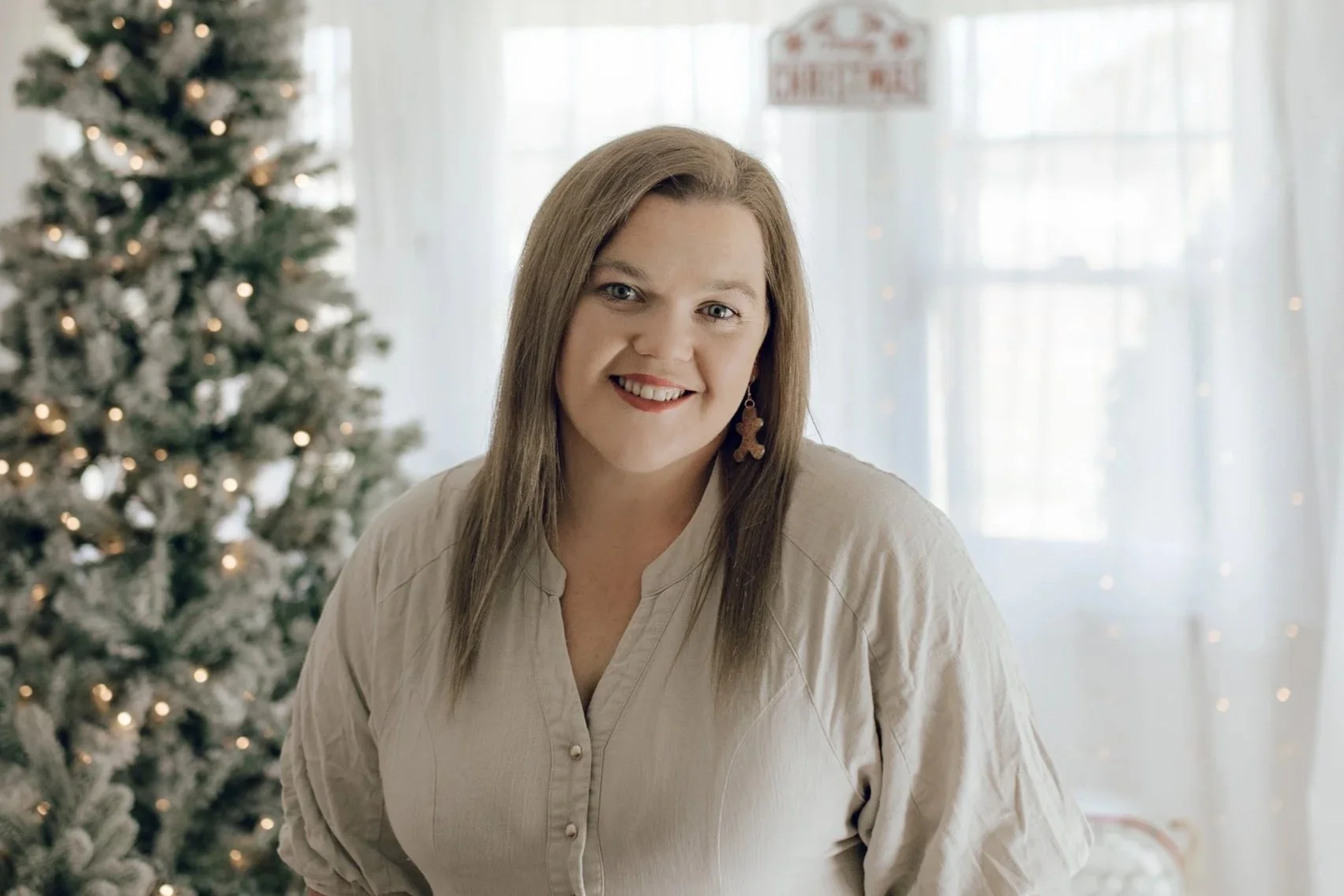 A woman with long brown hair, wearing a beige blouse and gingerbread man earrings, smiling in front of a Christmas tree decorated with lights, with a window and sheer curtains in the background.