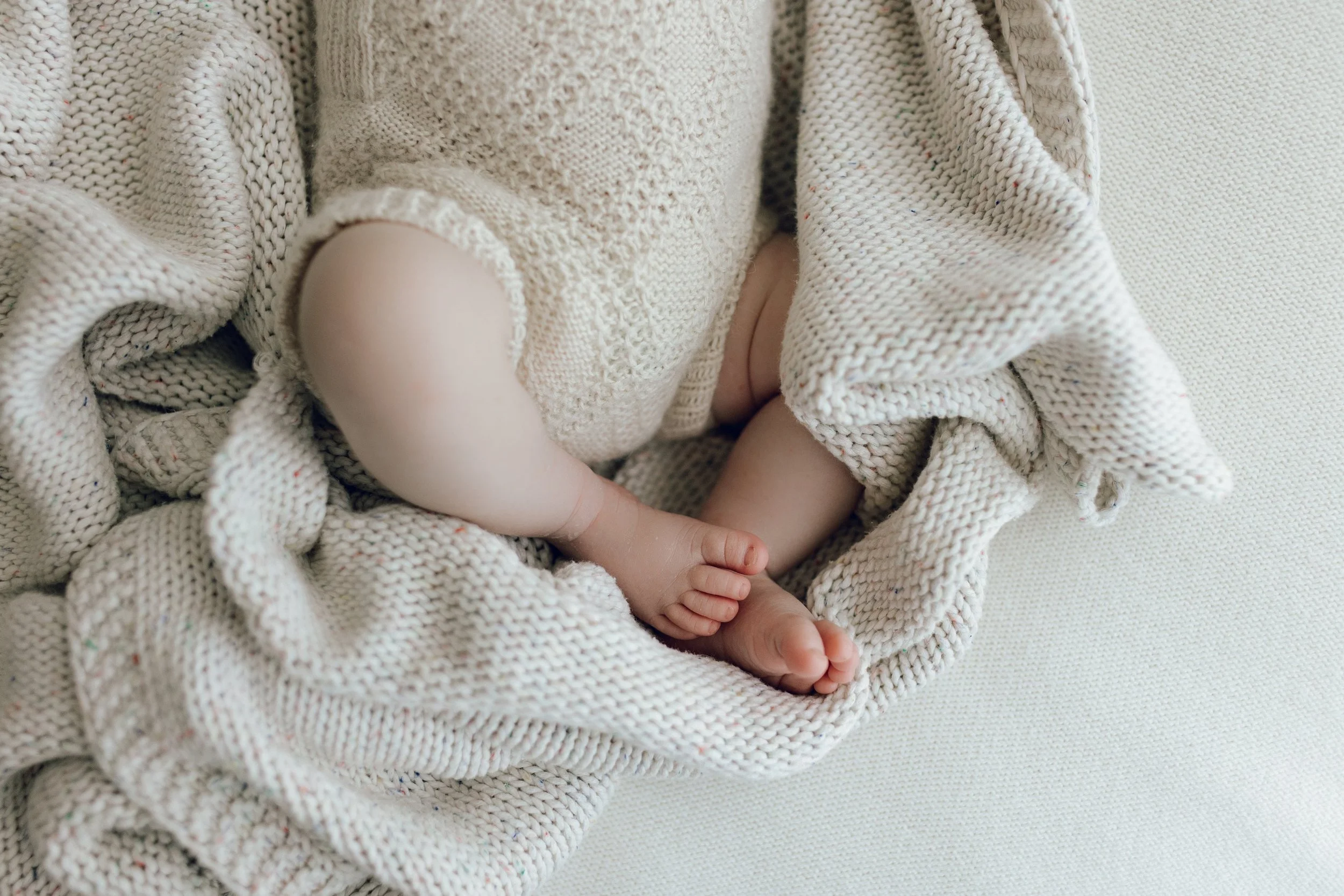 Close-up of a baby’s legs and feet, wrapped in a cream-colored knitted blanket, lying on a light-colored surface.