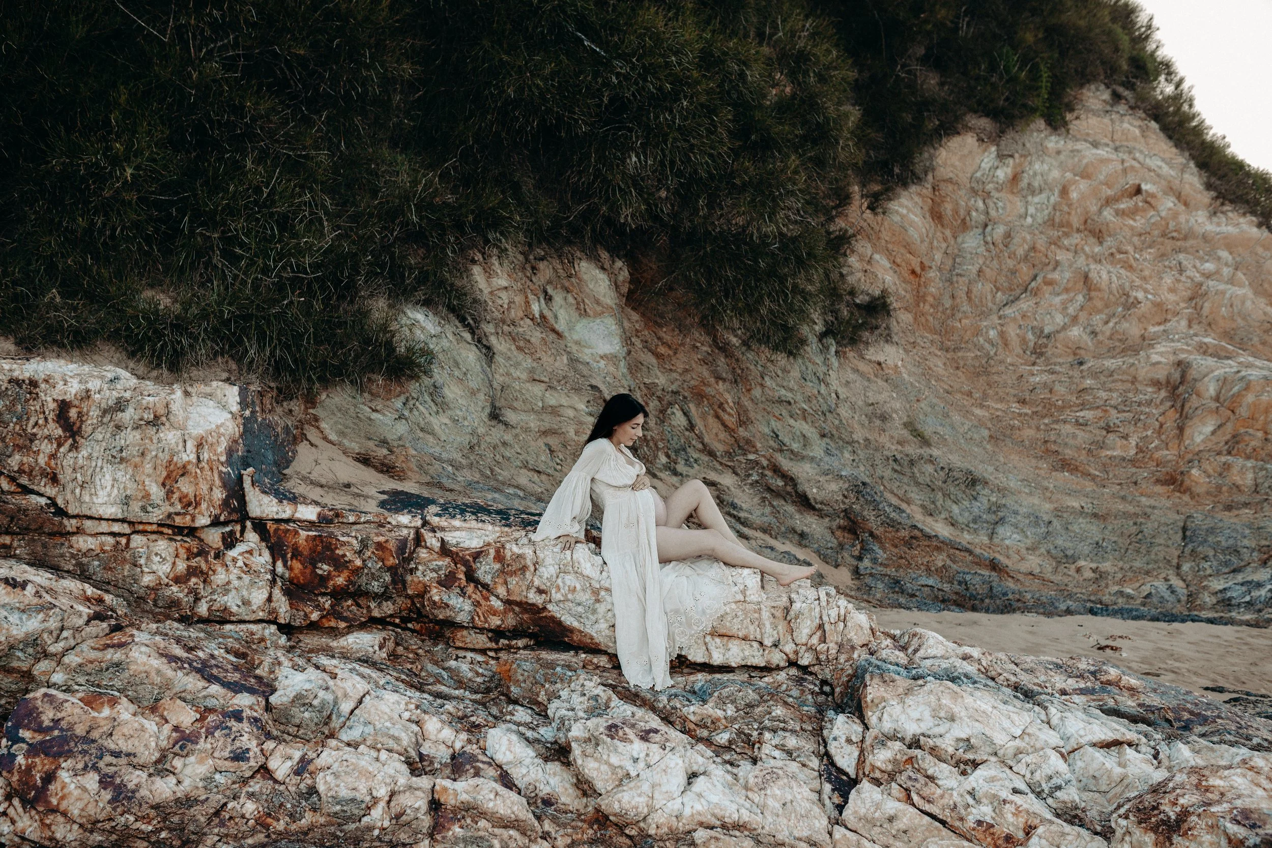 A woman in a white dress sitting on rocks at the beach near a rocky hillside with green foliage.