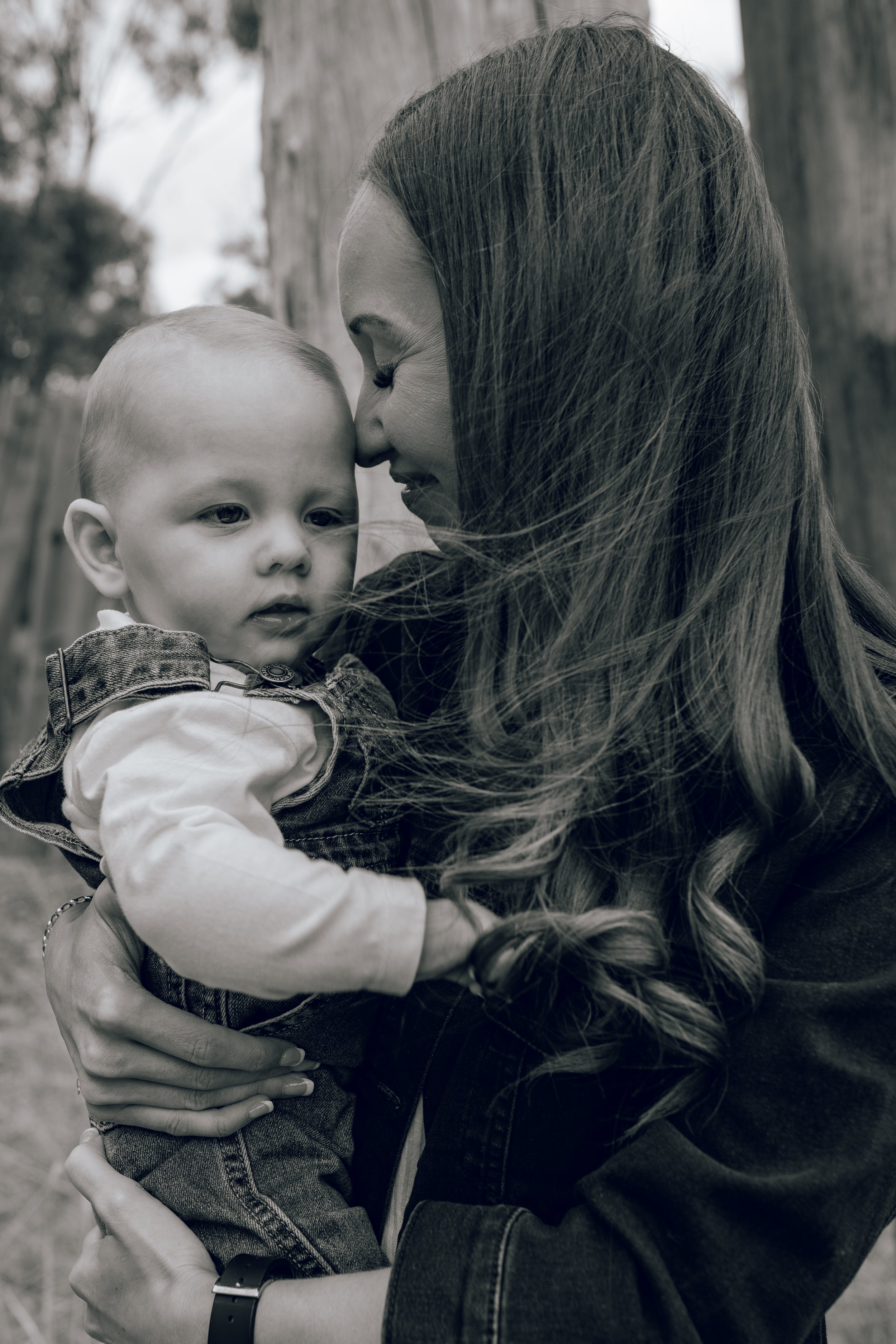 A woman holding a young child closely with their foreheads touching in an outdoor setting.