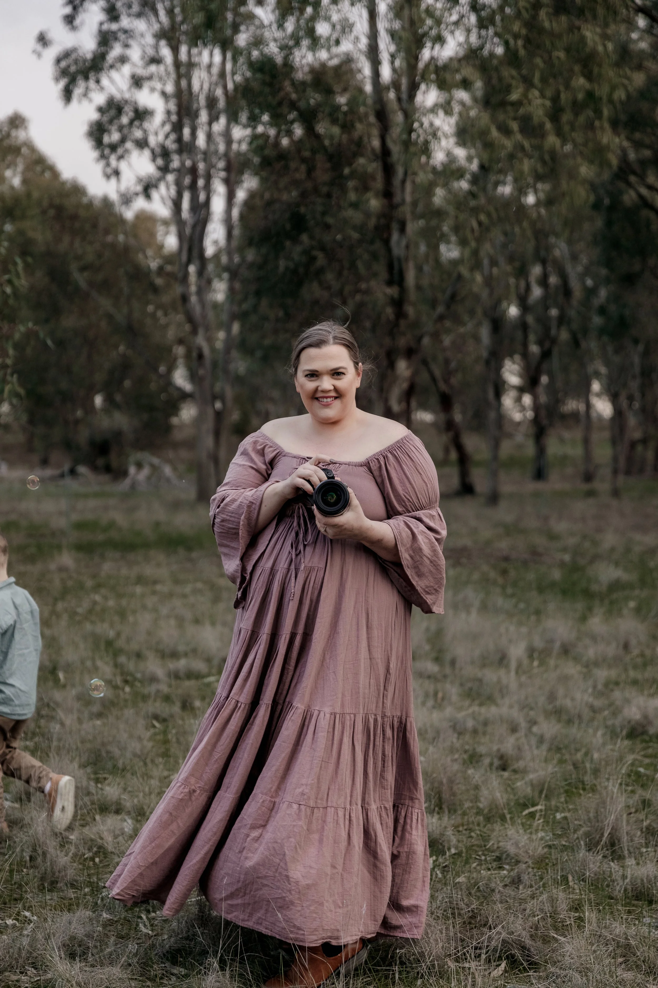 A woman in a mauve off-the-shoulder maxi dress holding a camera in an outdoor setting with trees and grass.