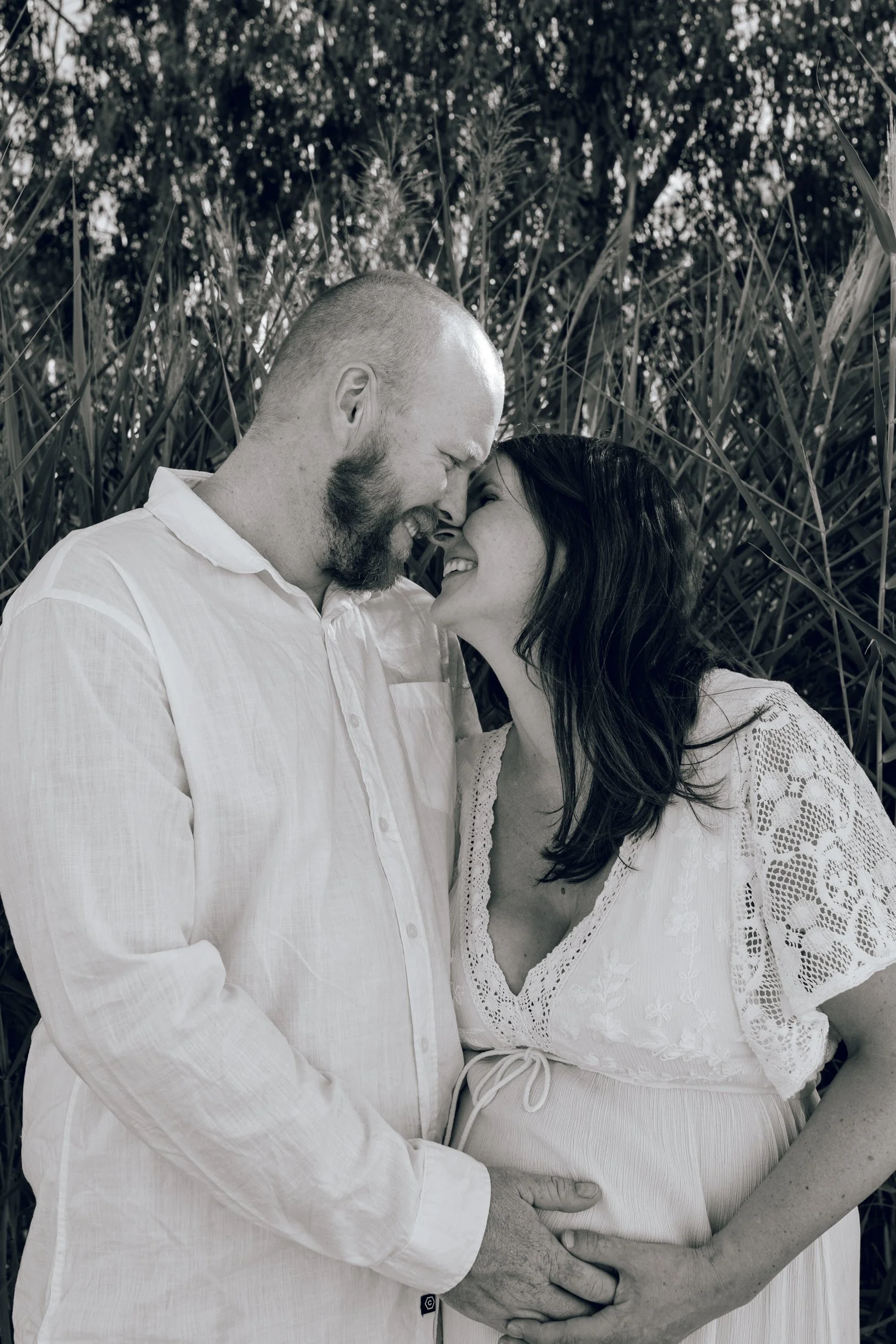 A black and white photo of a couple smiling and touching foreheads, standing close together outdoors with tall grass in the background.
