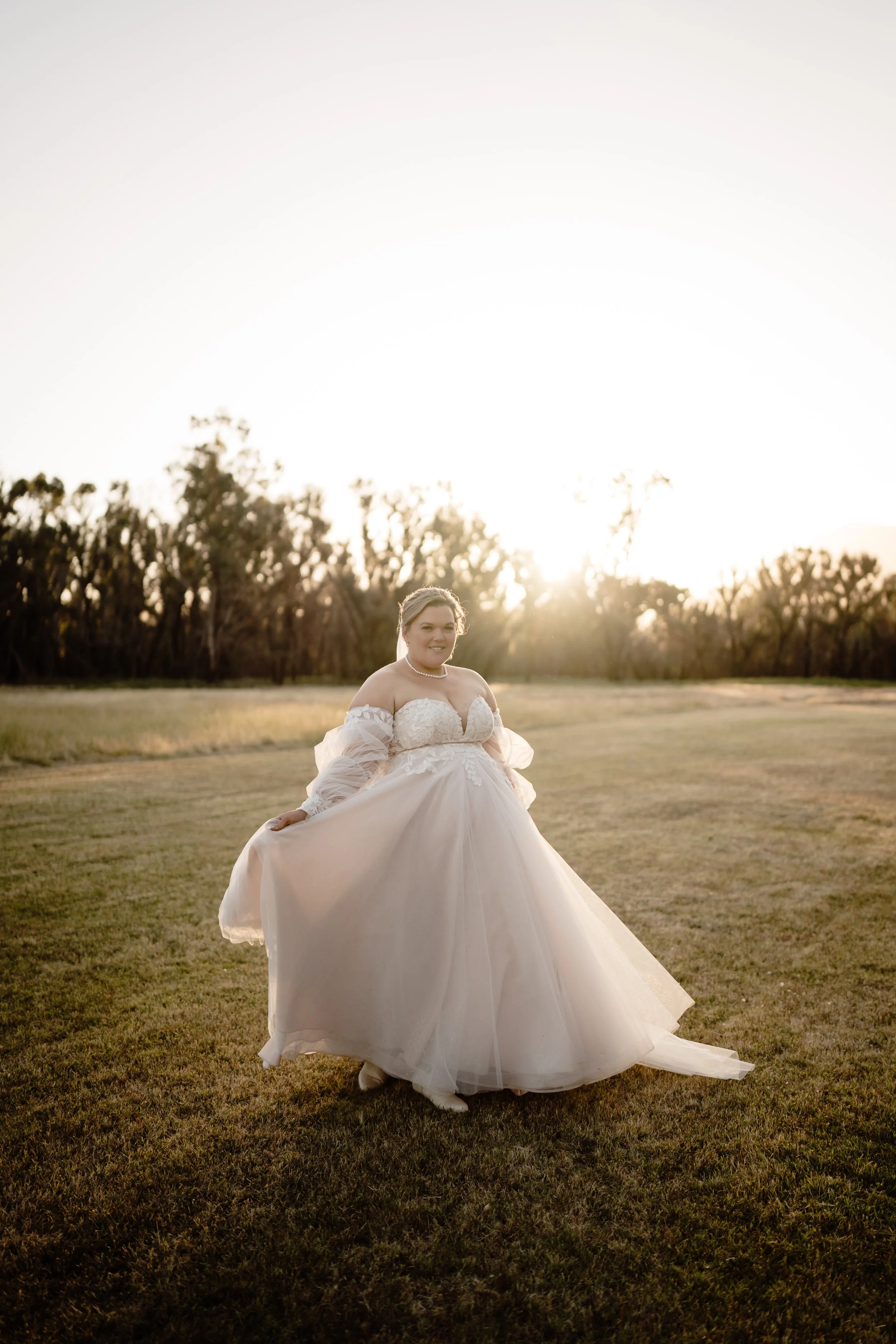 A woman in a wedding dress standing on a grassy field during sunset.