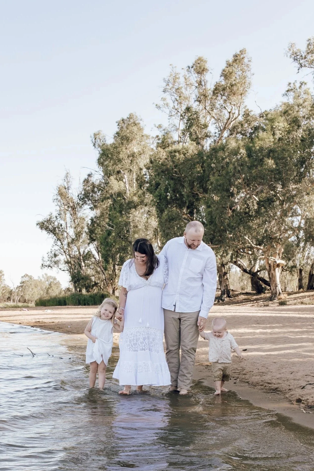 A family of four walking along the shoreline at the beach, holding hands and smiling, with trees in the background and clear sky.