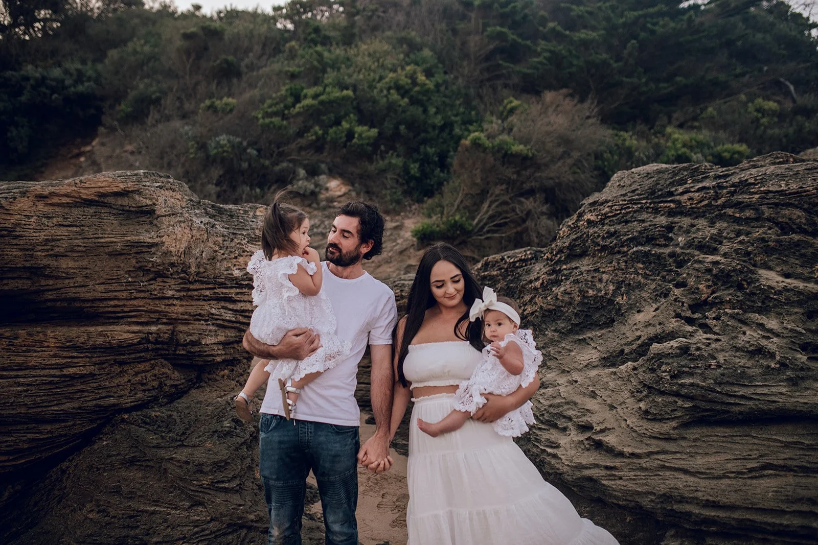 A family of four outdoors on a rocky area with trees in the background. The father is holding a young girl in a white dress, and the mother is holding a baby girl in a white dress with a large bow. The family is holding hands and looking at each other.