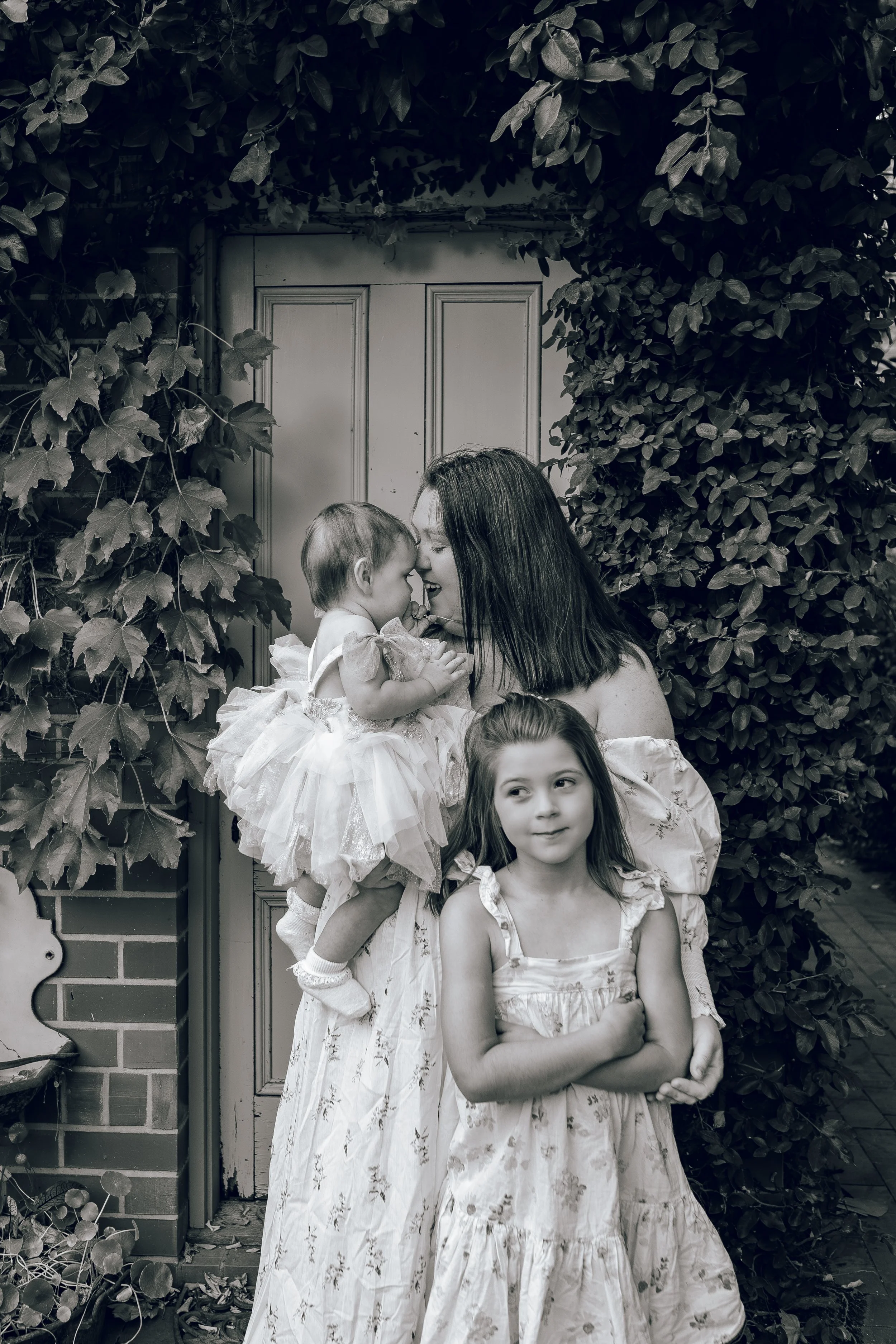 A woman with two young girls standing outside in front of a door surrounded by foliage, with one girl sitting on the woman's shoulder and the other girl standing in front with arms crossed.