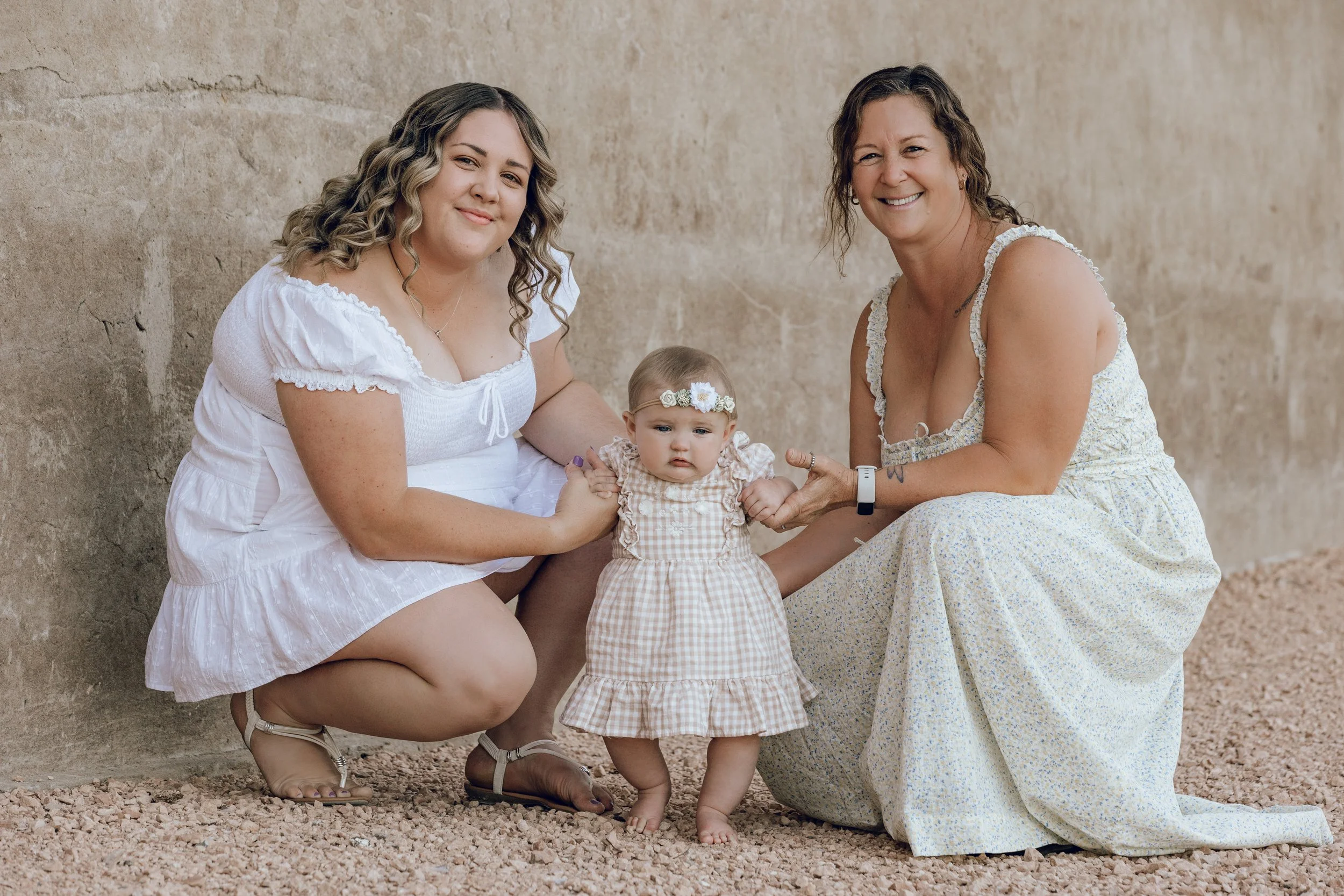Two women and a young girl pose outdoors in front of a stone wall. The women are kneeling and holding the girl, who wears a checkered dress and a floral headband. Everyone is smiling.
