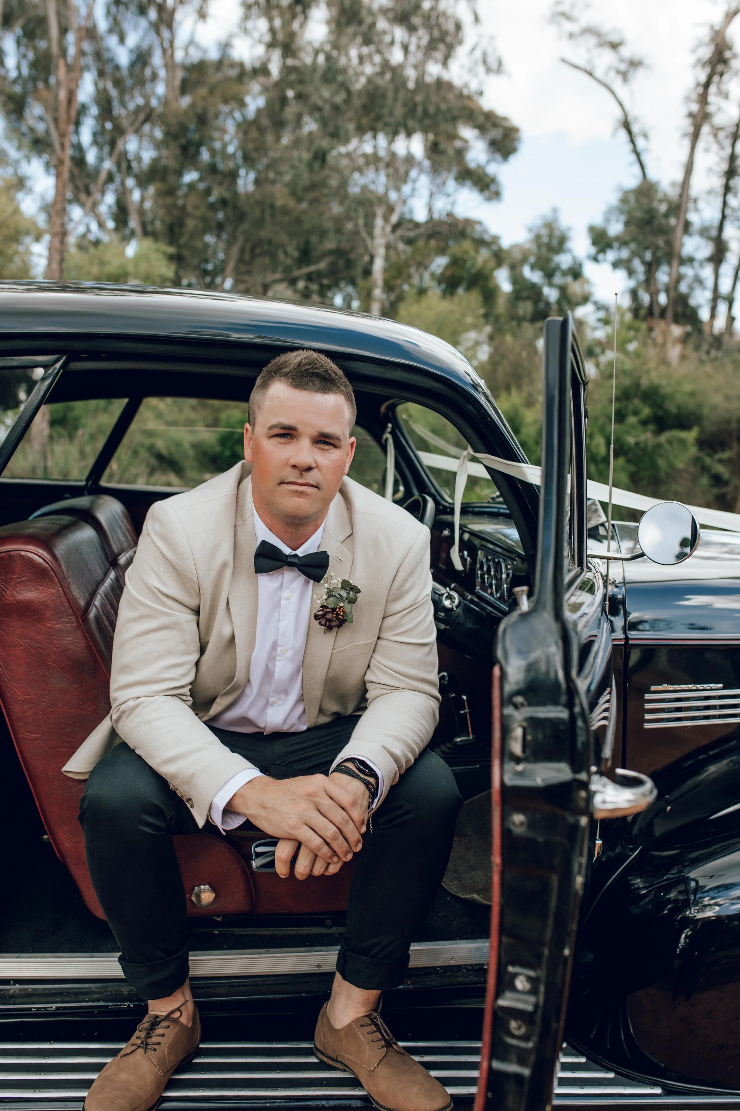 A man dressed in a beige blazer, white shirt, black bow tie, and dark pants sitting in a vintage black car with a red interior, outdoors with trees in the background.