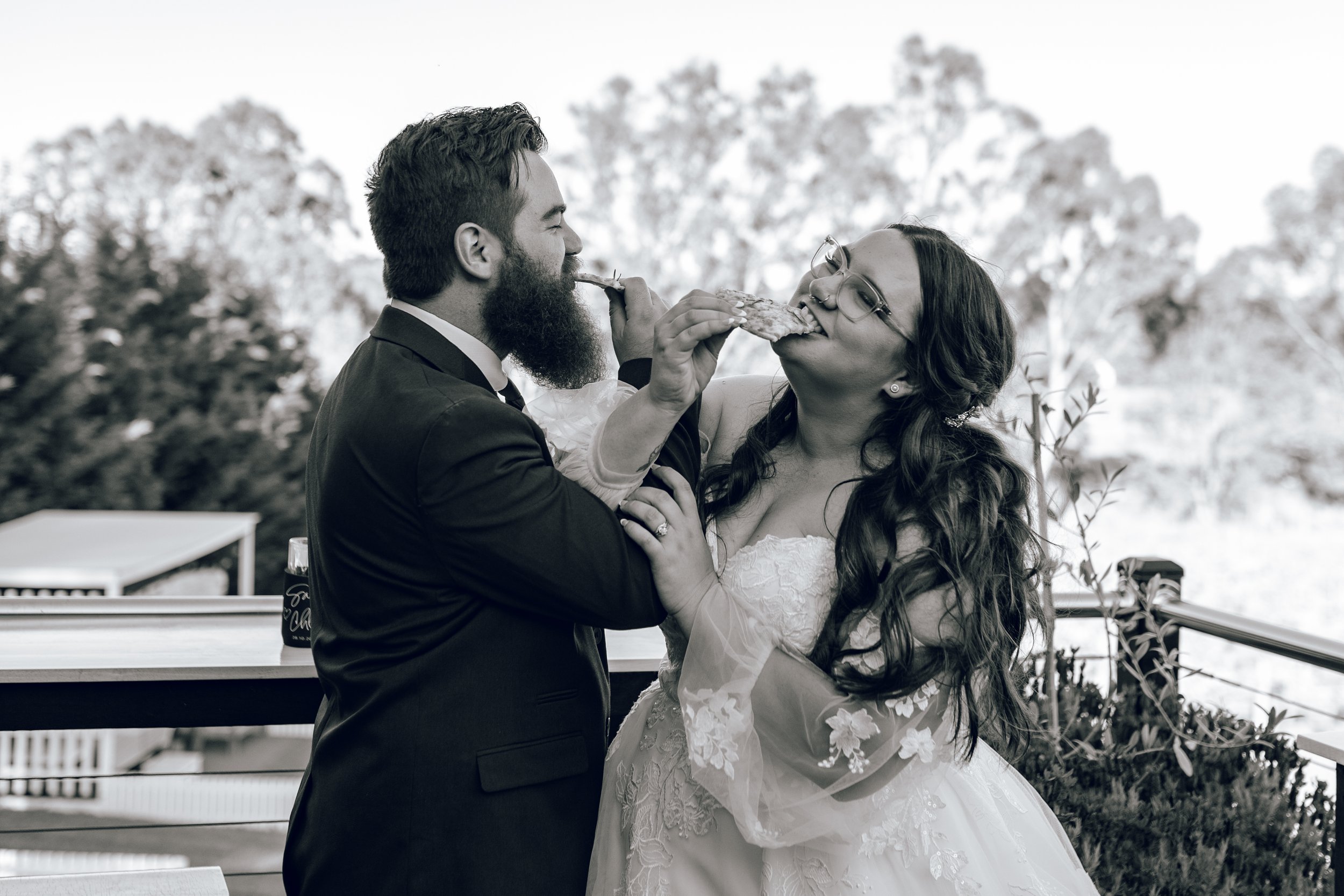 A black-and-white photo of a bride and groom sharing a slice of pizza during their wedding celebration outdoors, with trees in the background.