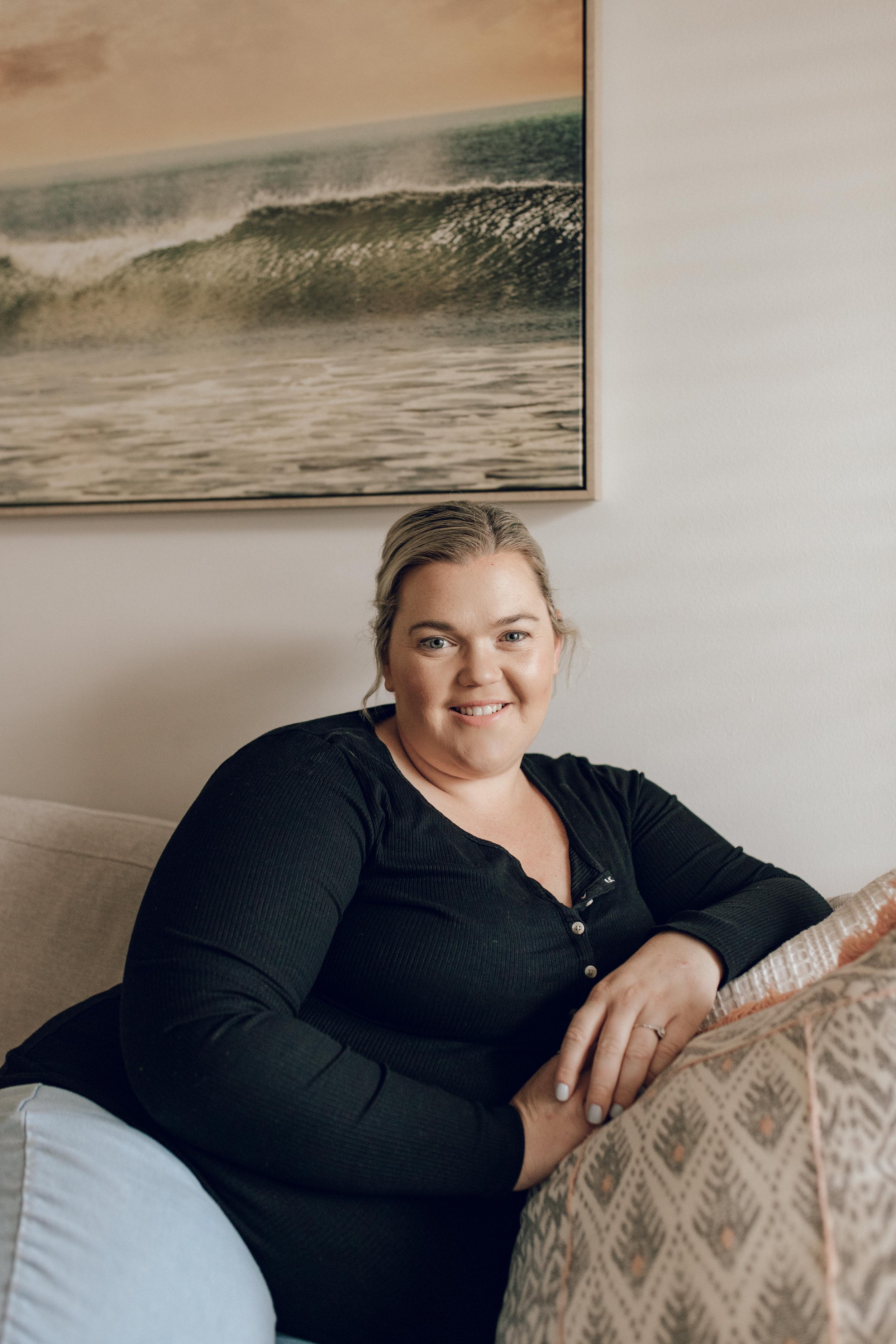 A smiling woman in a black top sitting on a beige couch, with a patterned cushion in front of her, in a room with a large painting of ocean waves on the wall behind her.