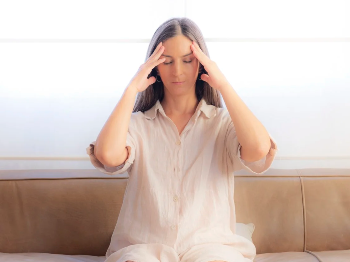 A woman sitting on a couch with her eyes closed, hands pressed against her temples, appearing to aleviate a headache or stress. Somatic touch. migranes and headaches.