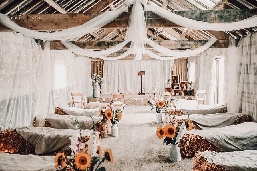 Interior of a rustic barn with two rows of beds covered in burlap, string lights hanging from the exposed wooden beams, and sheer curtains along the sides.