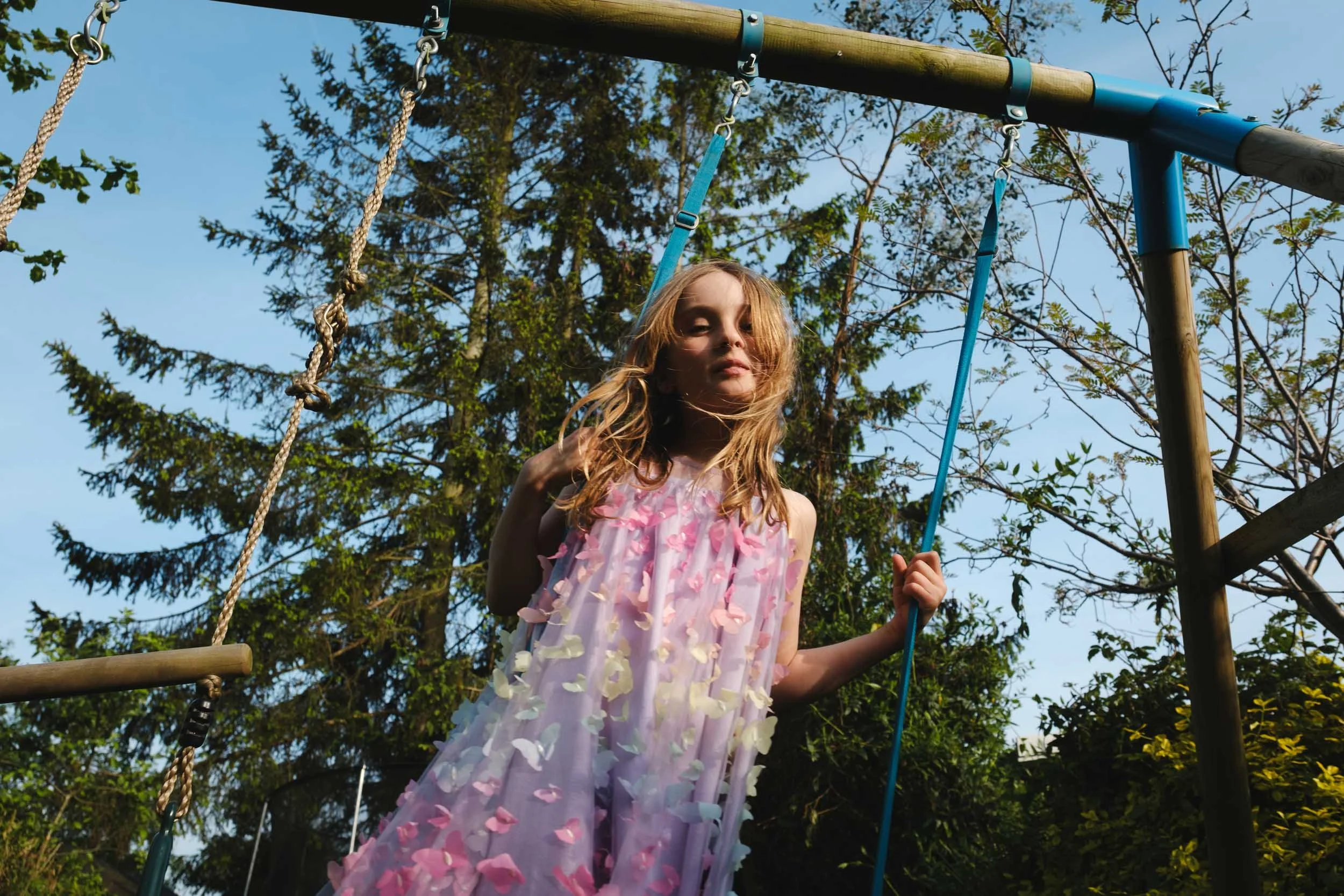 A young girl with long blonde hair wearing a pink dress with flower appliques, standing on a playground swing set surrounded by trees and blue sky.