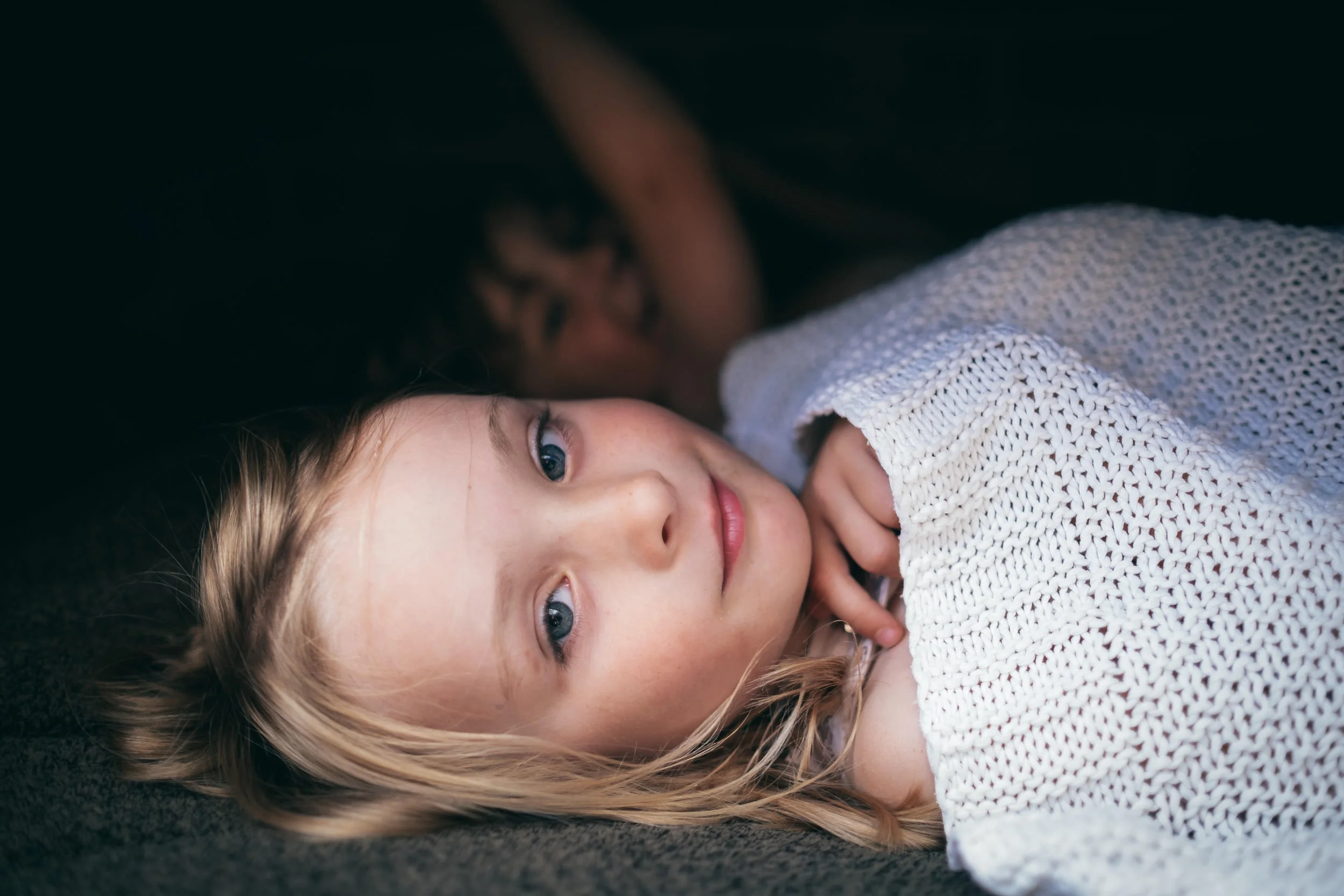 A young girl with blonde hair and blue eyes lying on a dark surface, wearing a cream-colored knit sweater, looking into the camera with a gentle expression.