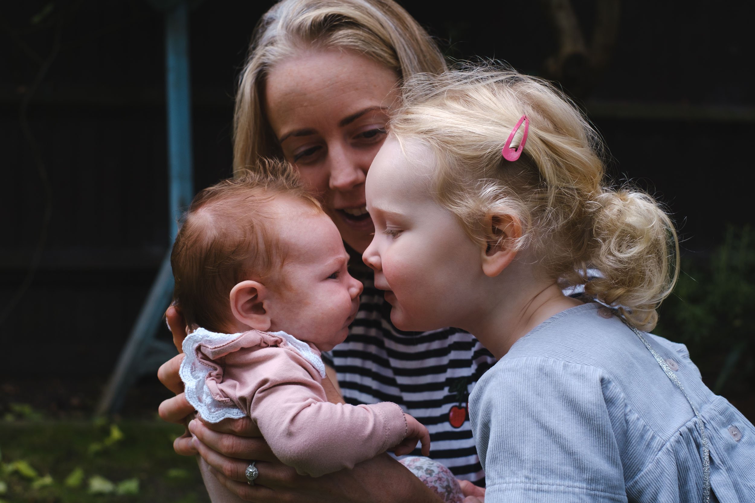 A woman with blonde hair and a young girl with curly blonde hair, wearing a pink hair clip, are touching foreheads and smiling at a baby girl with red hair, in an outdoor setting.