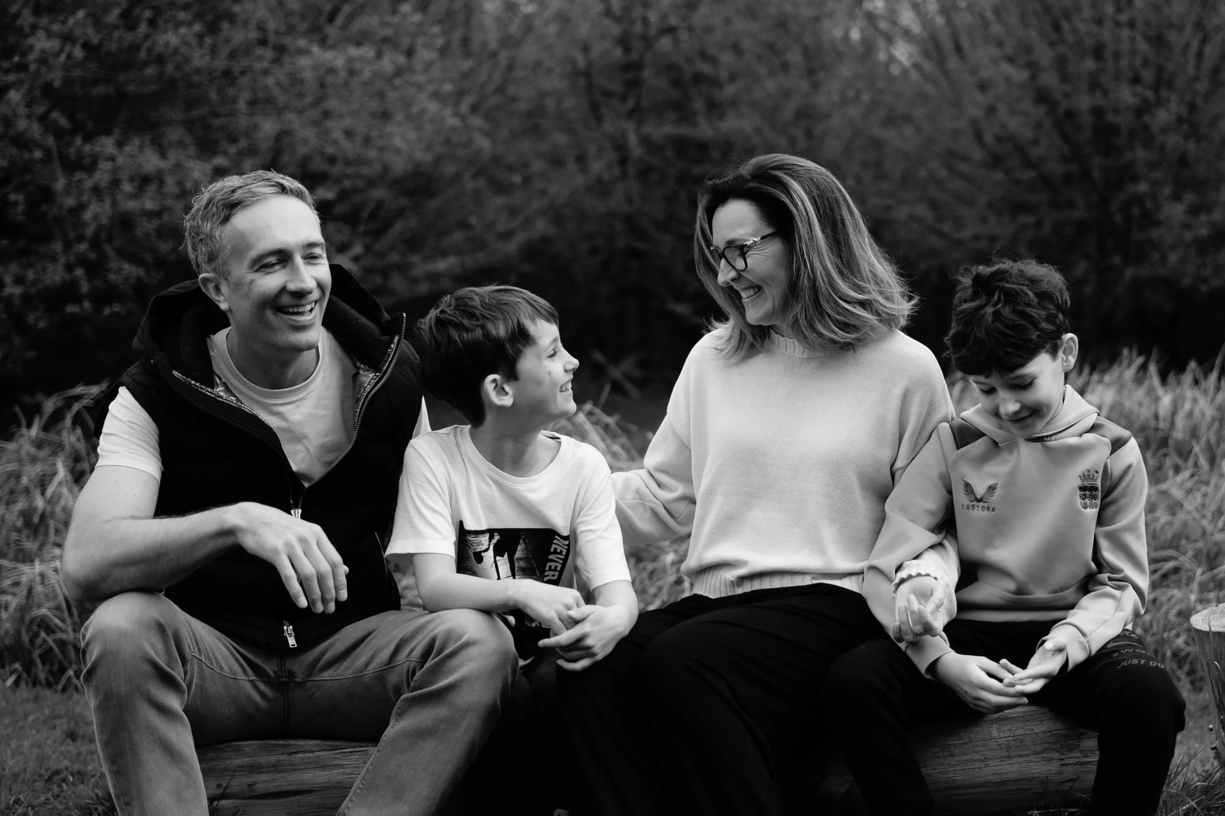 A black and white photograph of a smiling family of five sitting outdoors on a large log, with a background of trees and grass. They are engaging in a cheerful conversation.