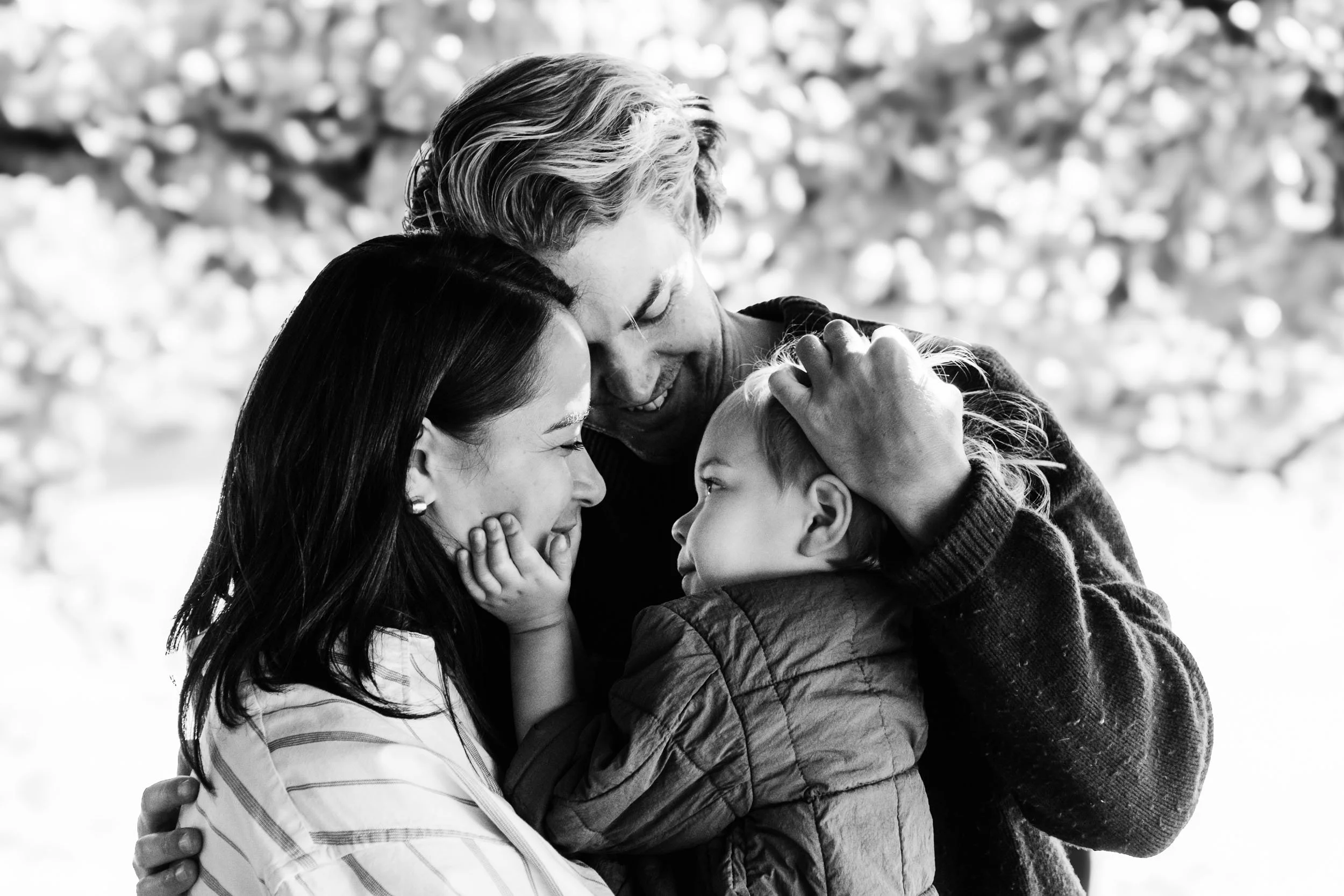 Black and white photo of a family embracing outdoors with a background of blurred trees and sunlight.