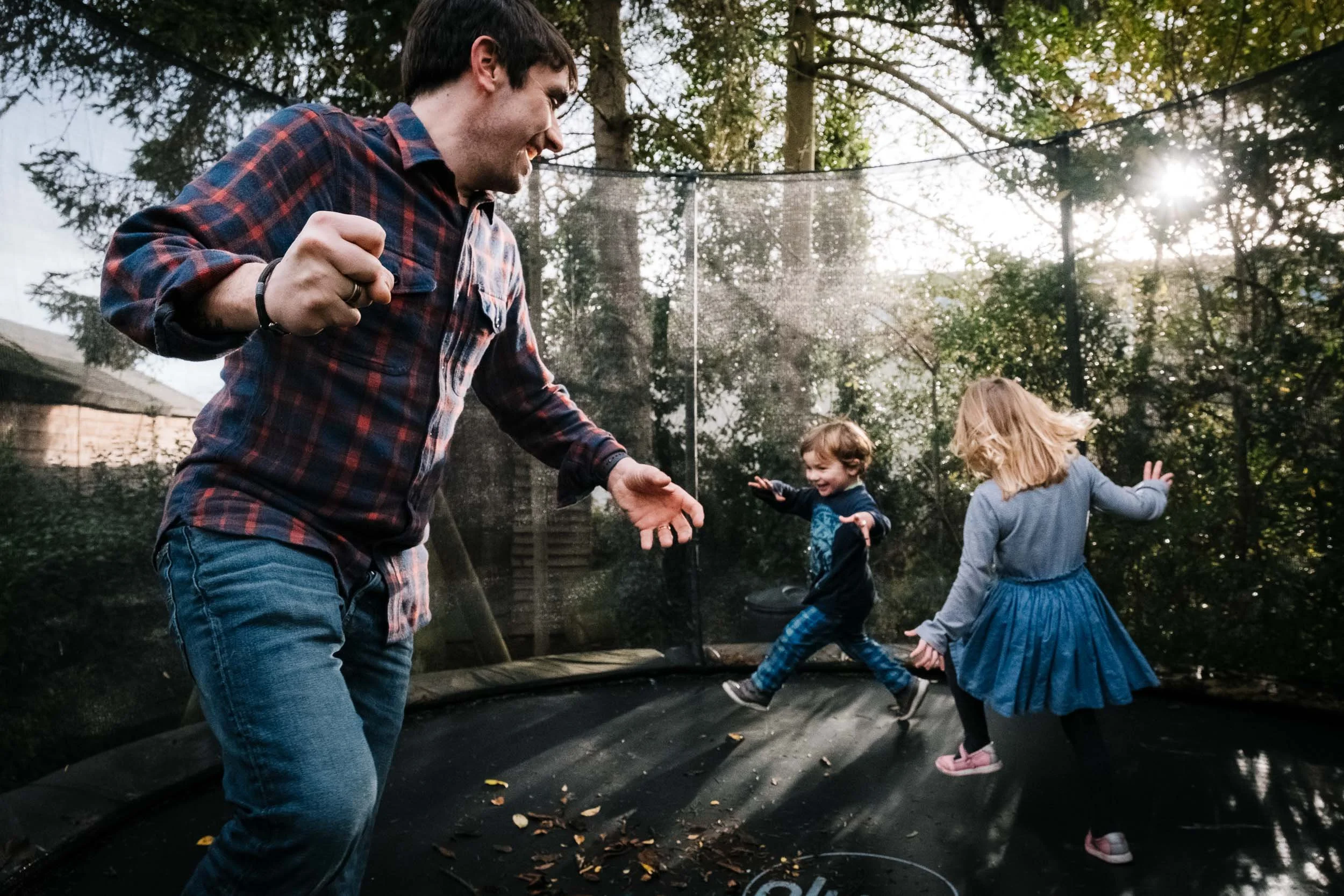 A man and two young girls are playing and jumping on a trampoline outside in a wooded backyard during late afternoon or early evening, with sunlight shining through trees.