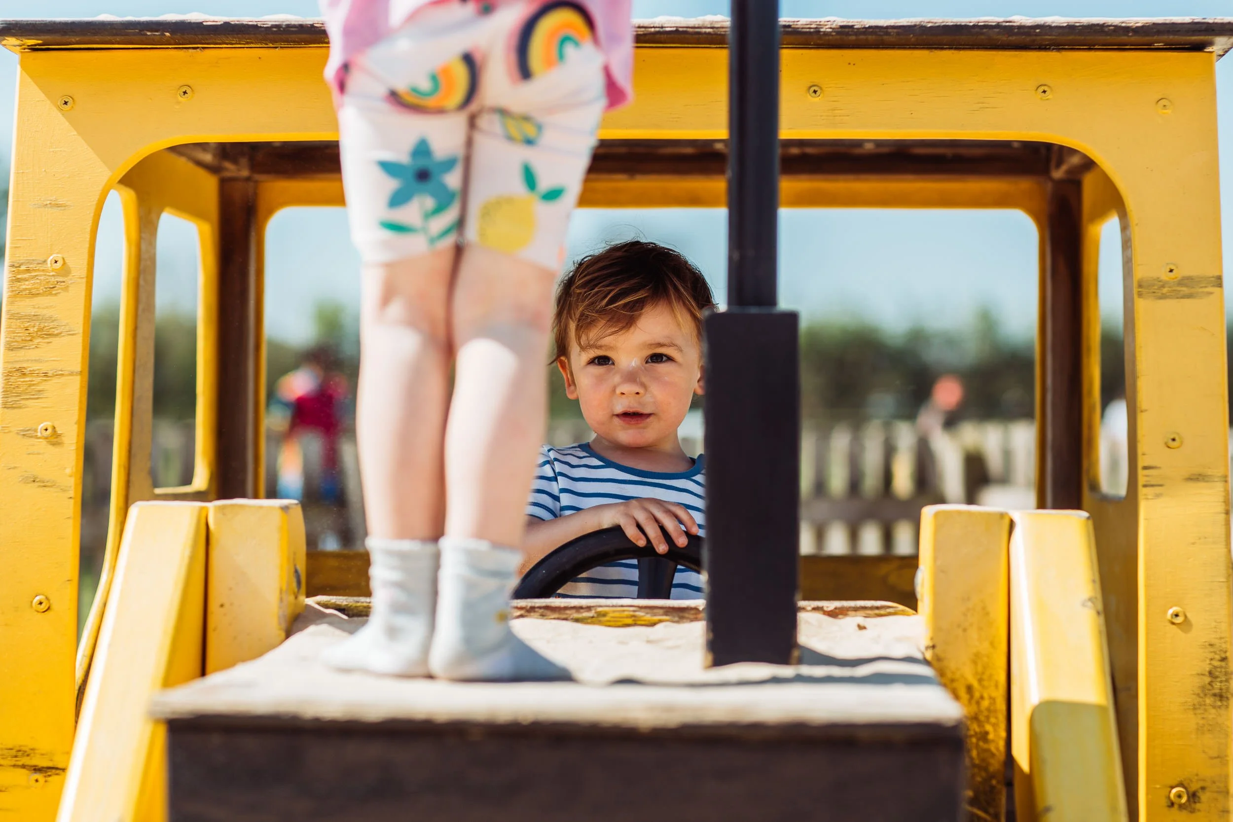 A young boy with brown hair and a striped blue and white shirt looking out from behind a yellow playground structure, with someone’s legs and rainbow-patterned socks visible in the foreground.