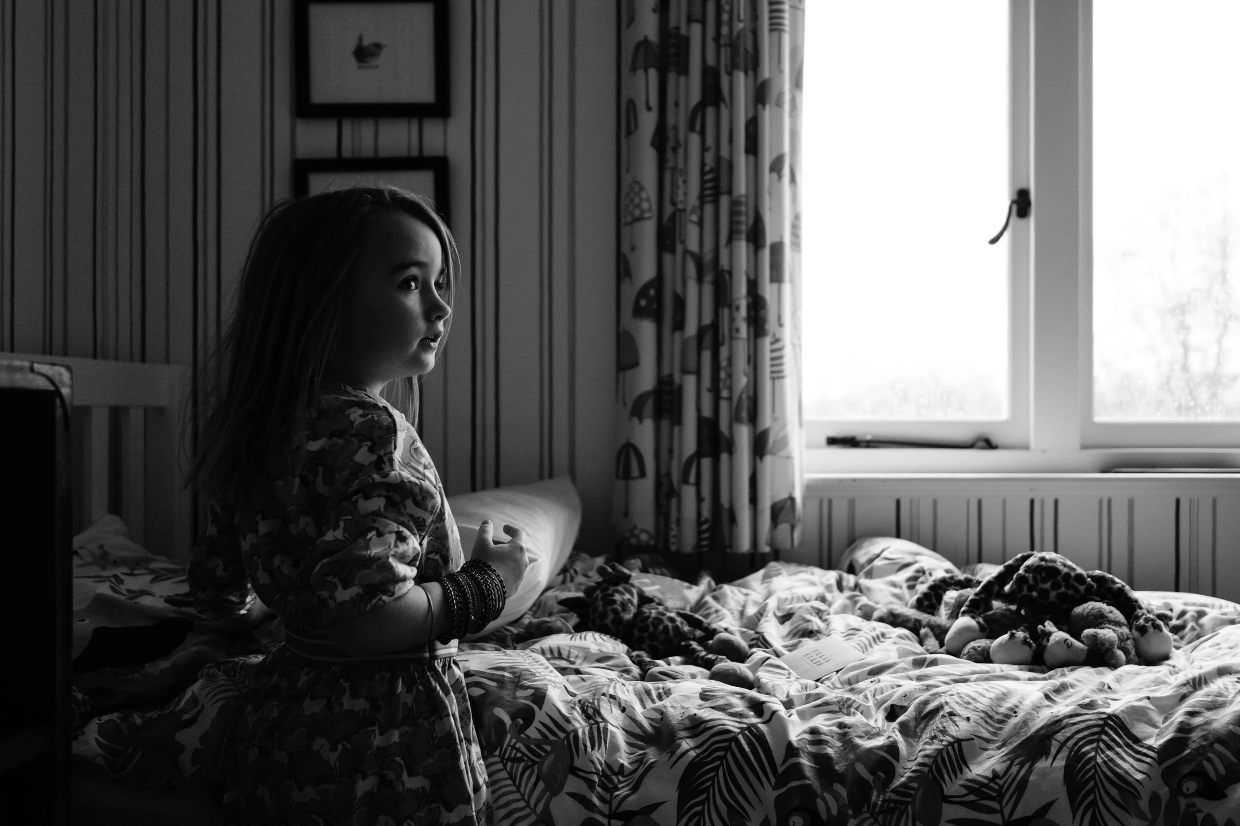 A young girl with long hair standing beside a bed with stuffed animals and pajamas, looking towards a window in a cozy bedroom.
