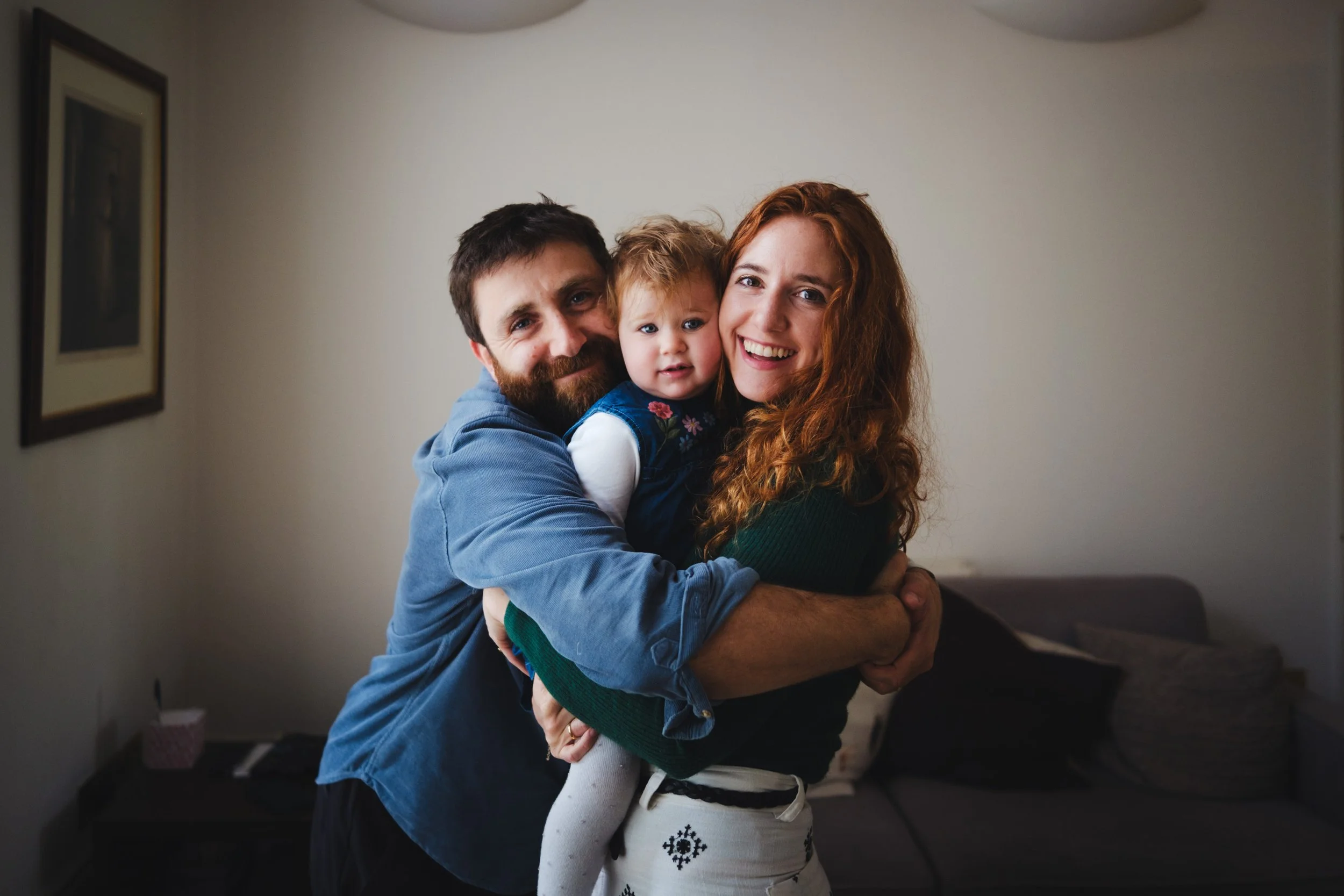A happy family of three hugging indoors, with a plain wall and framed artwork in the background.