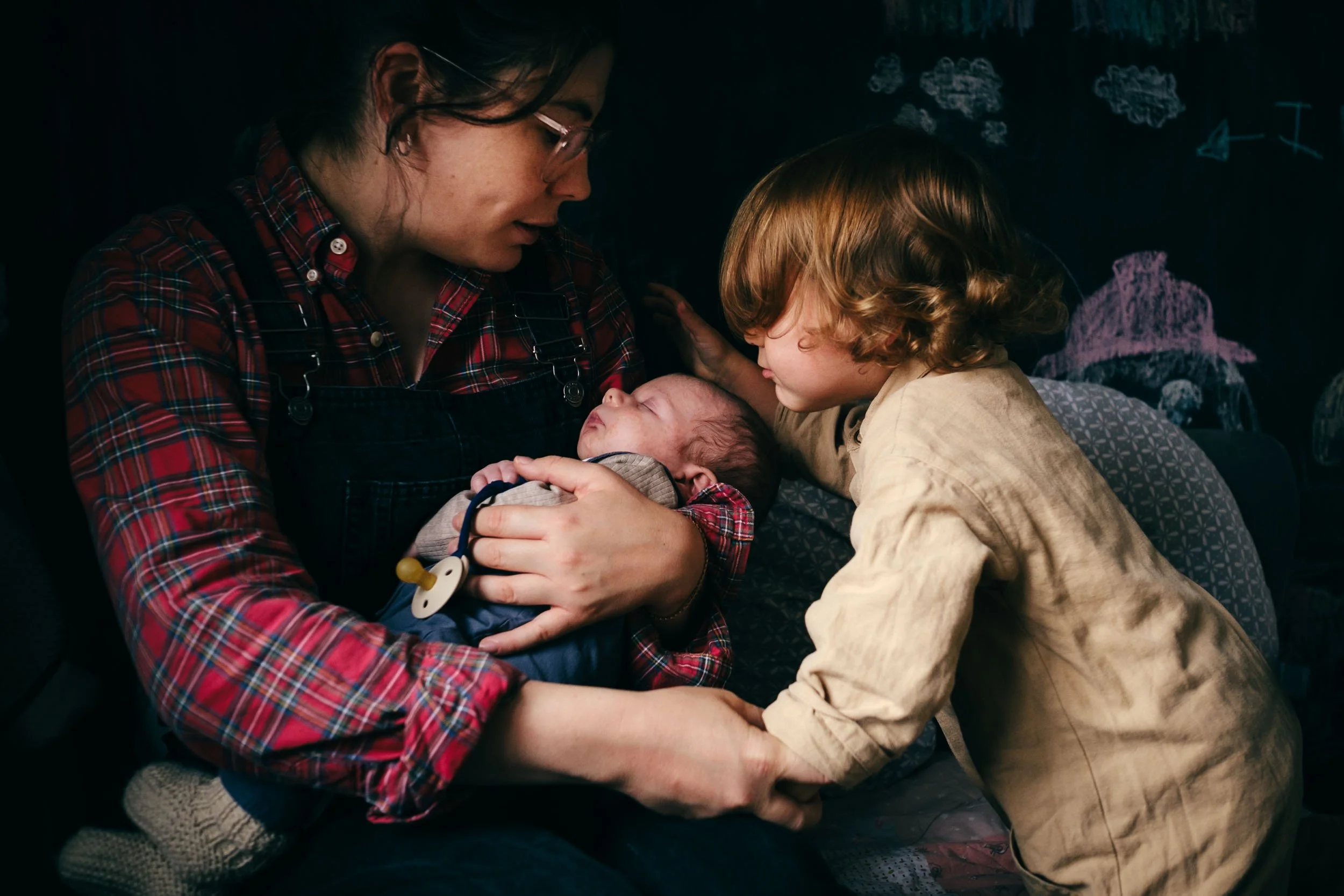 A woman and a young girl holding and looking at a newborn baby in a dimly lit room, with drawings of clouds and a mountain on a chalkboard in the background.