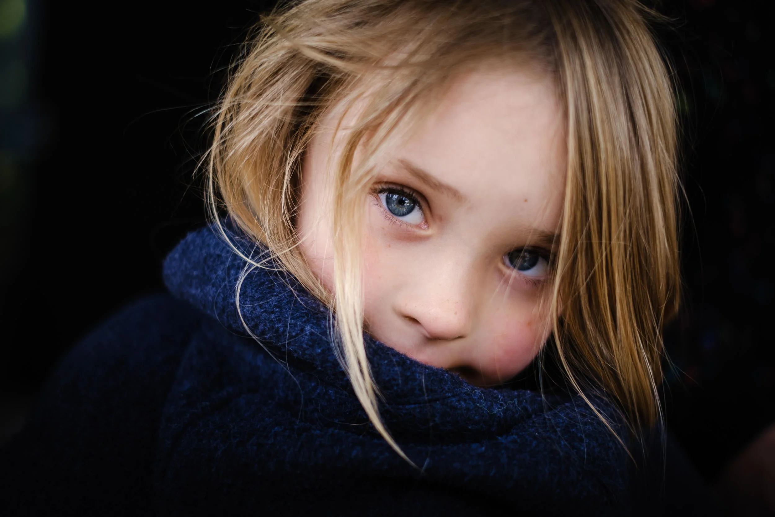 Close-up of a young girl with blue eyes and blonde hair, wearing a dark blue sweater, against a dark background.