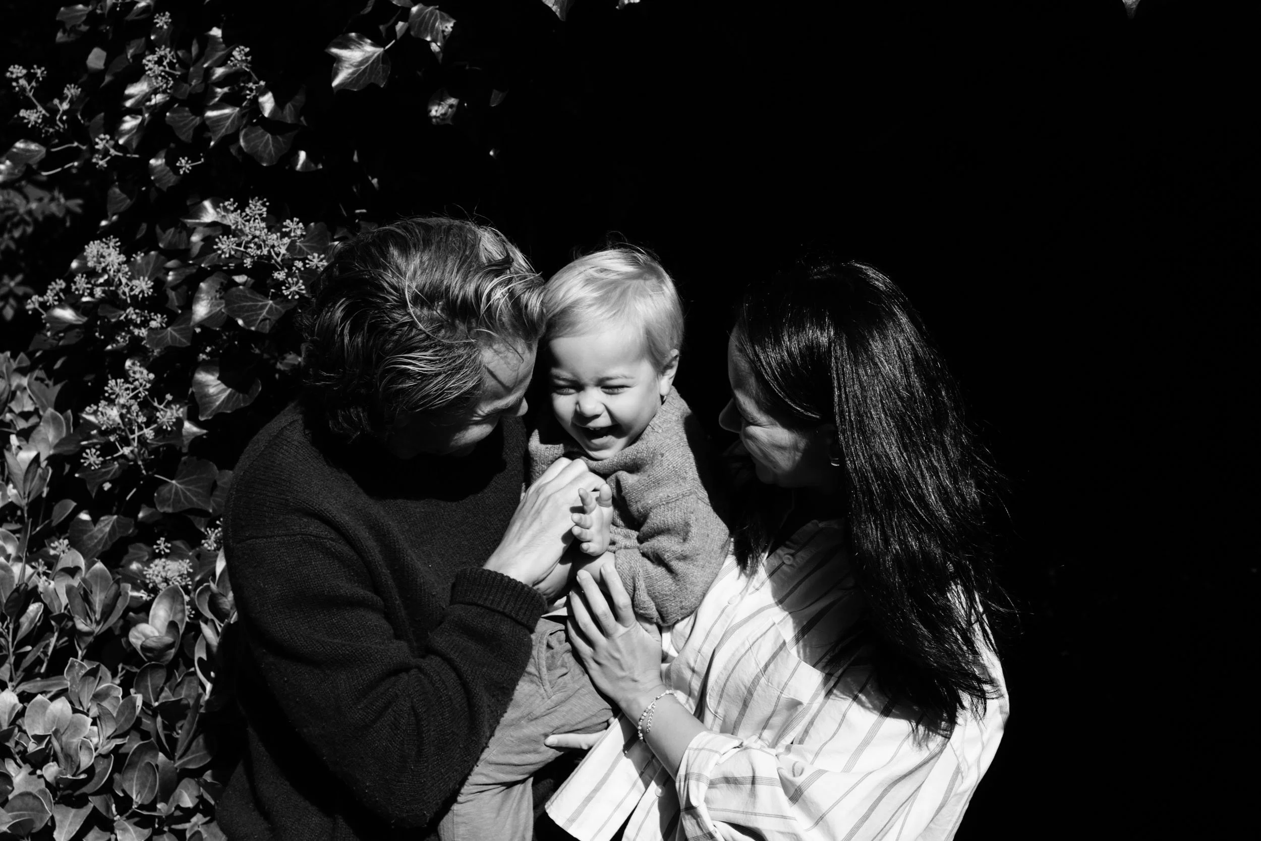 A joyful young boy is embraced by two women, one on each side, outside against a background of foliage. The boy is smiling and appears to be laughing, expressing happiness.