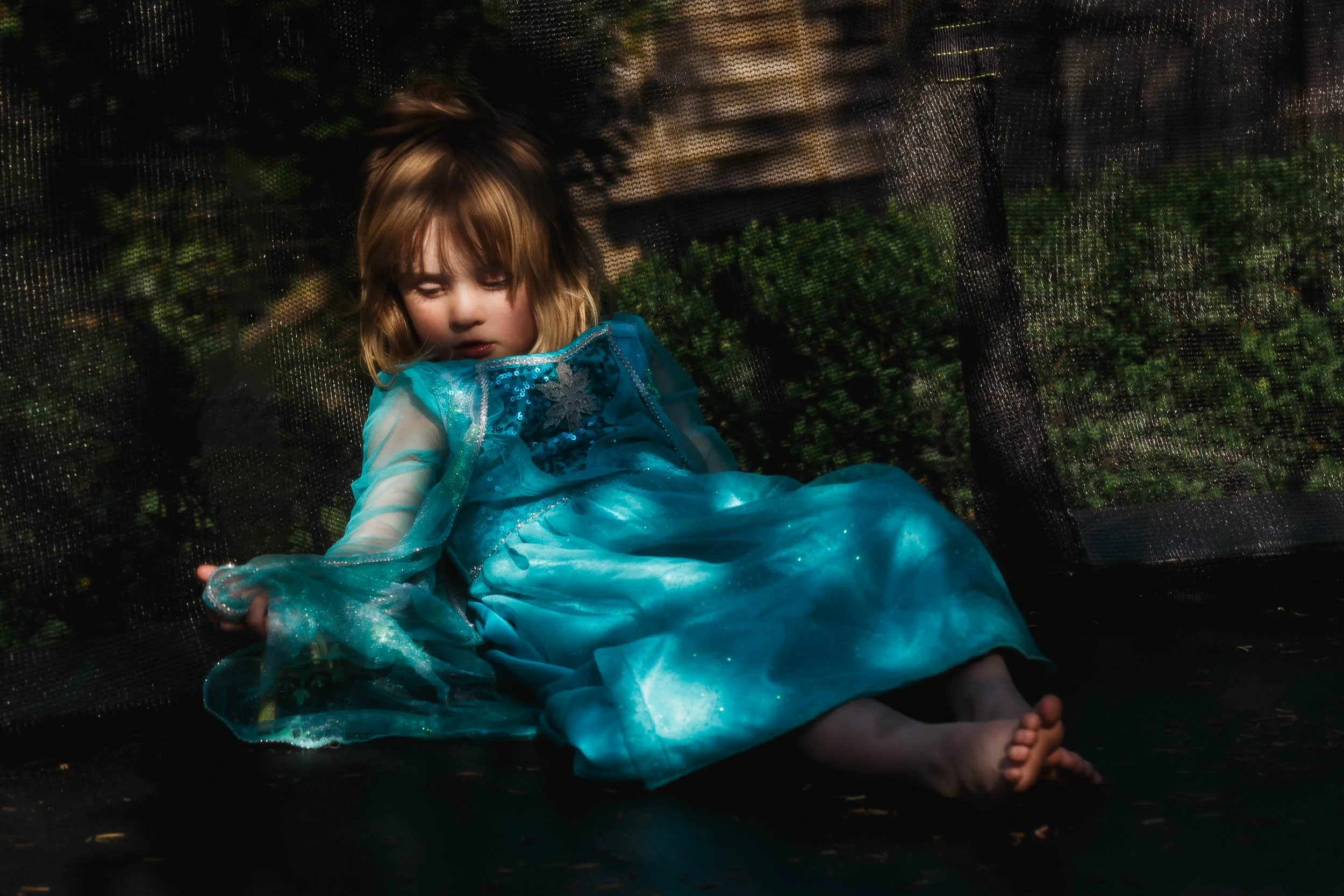 A young girl dressed as Elsa from Disney's Frozen, lying on a trampoline with her eyes closed, wearing a blue dress with sheer sleeves and snowflake embellishments, surrounded by greenery in the background.