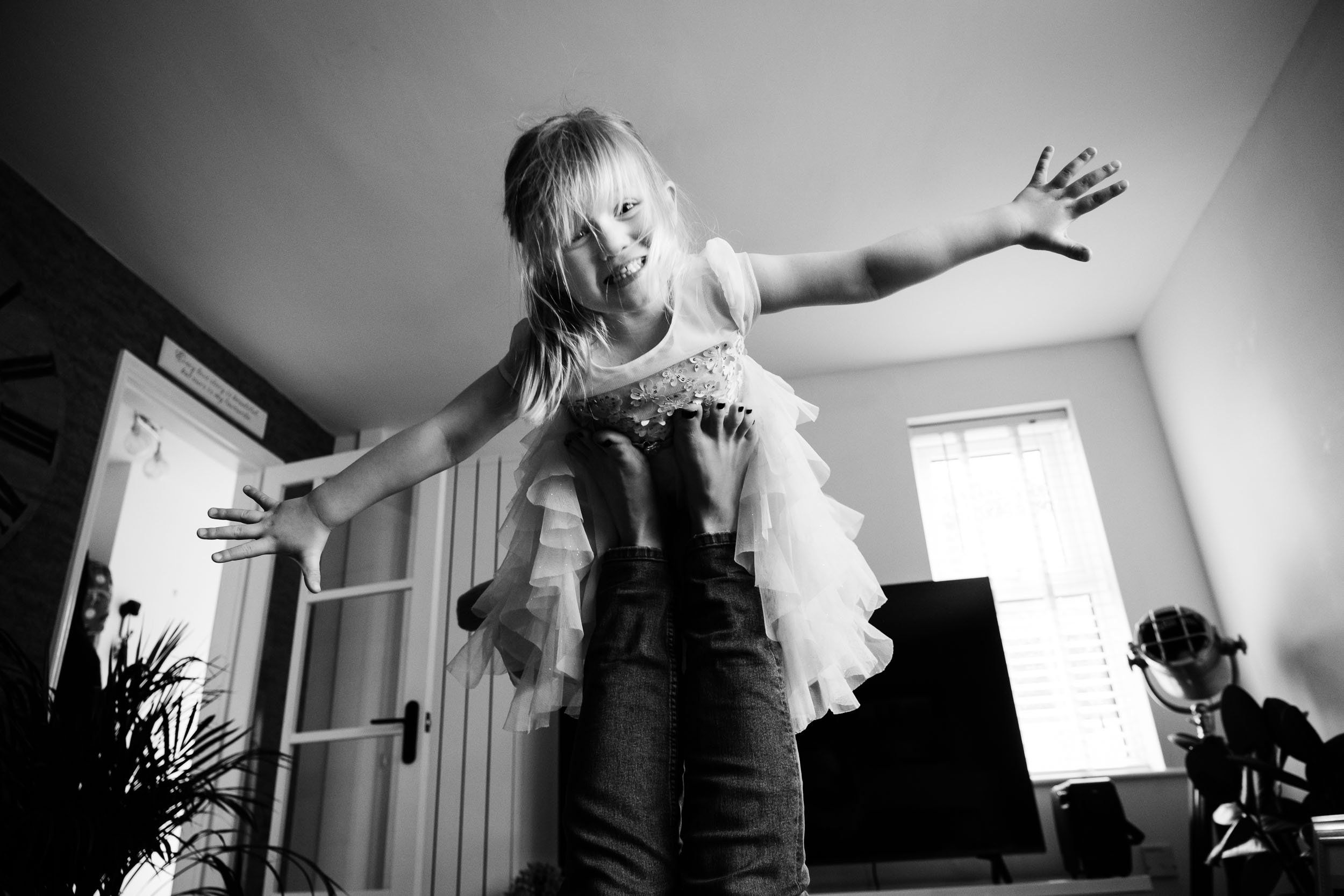 A young girl with long hair and a dress being held up by someone, smiling and extending her arms outward in a playful pose inside a room.