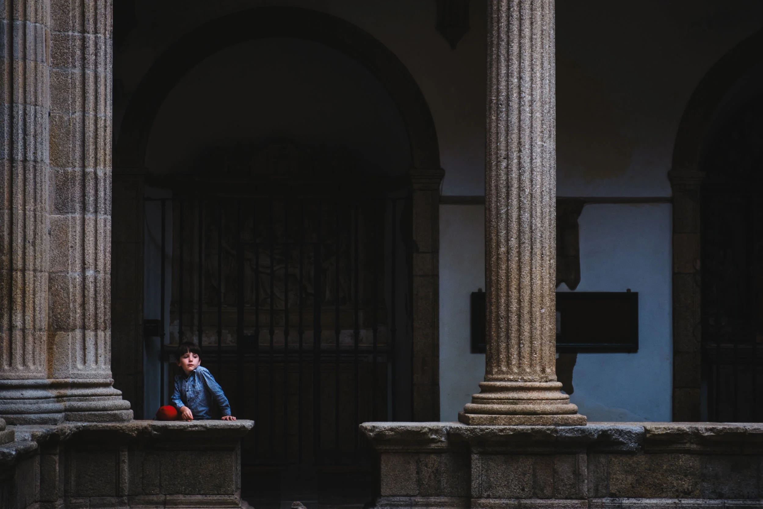 A young boy in a blue checkered shirt and red pants sitting on a stone ledge between two stone columns in a historical or architectural setting with arches and dark interior.