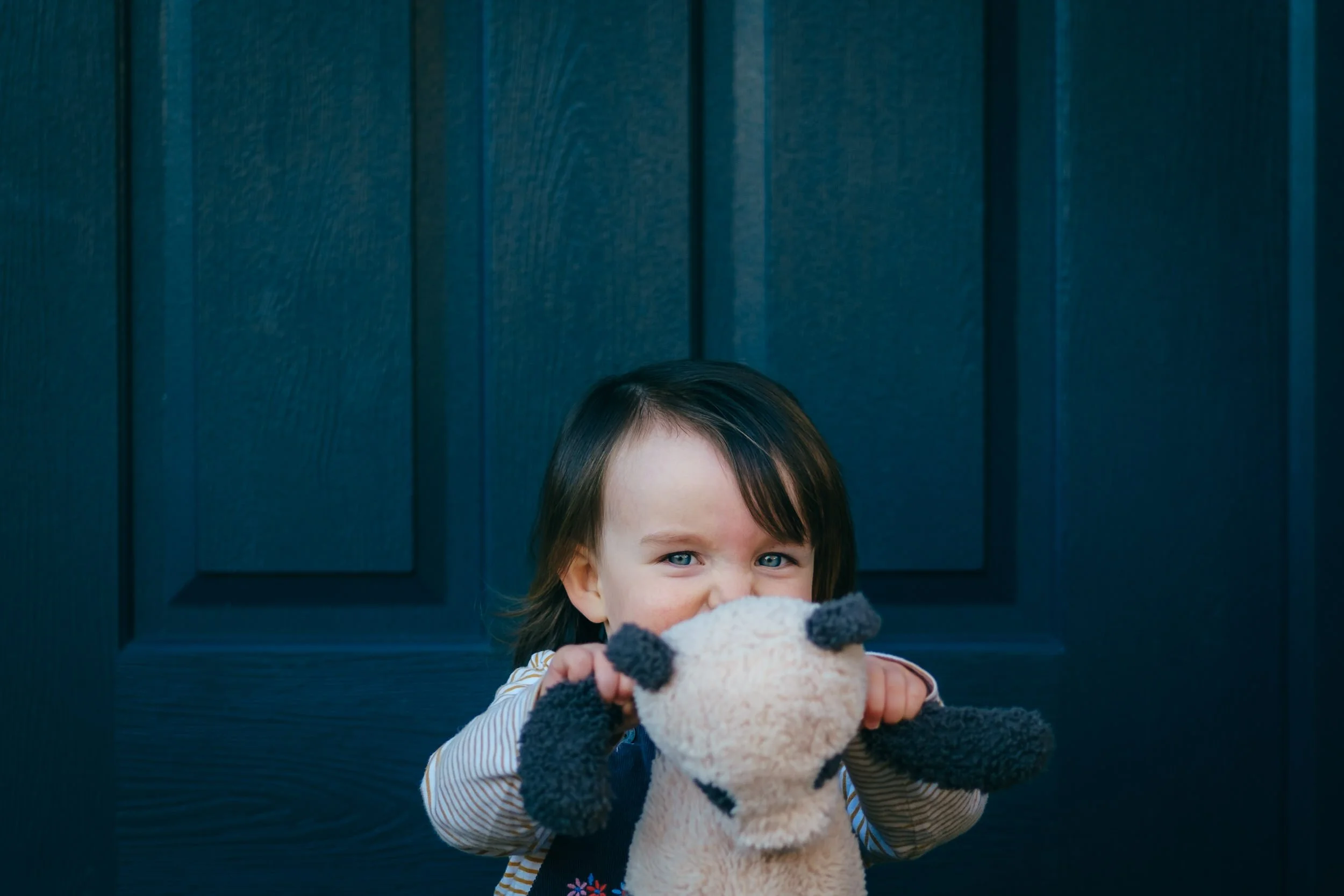 A young girl with brown hair and blue eyes holding a plush toy in front of her face, standing in front of a dark blue panelled front door.