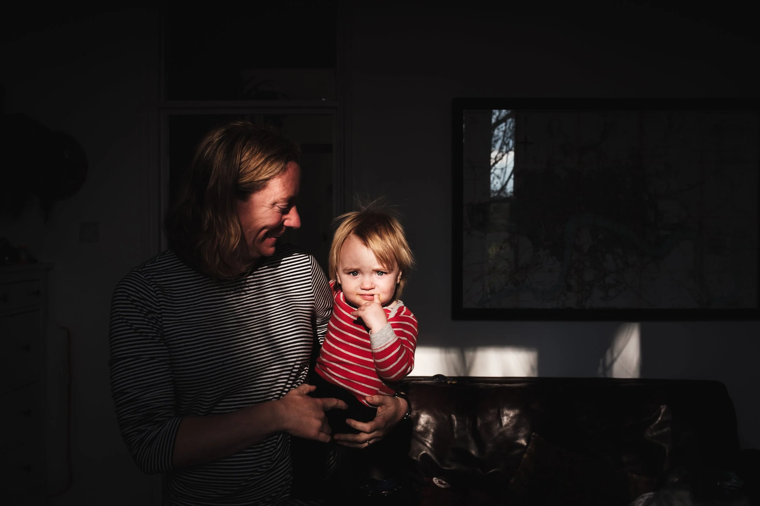 A woman with wavy hair and a striped shirt holding a young child in a striped sweater, both are in a dark room with a window in the background. The woman is smiling while the child looks curious.