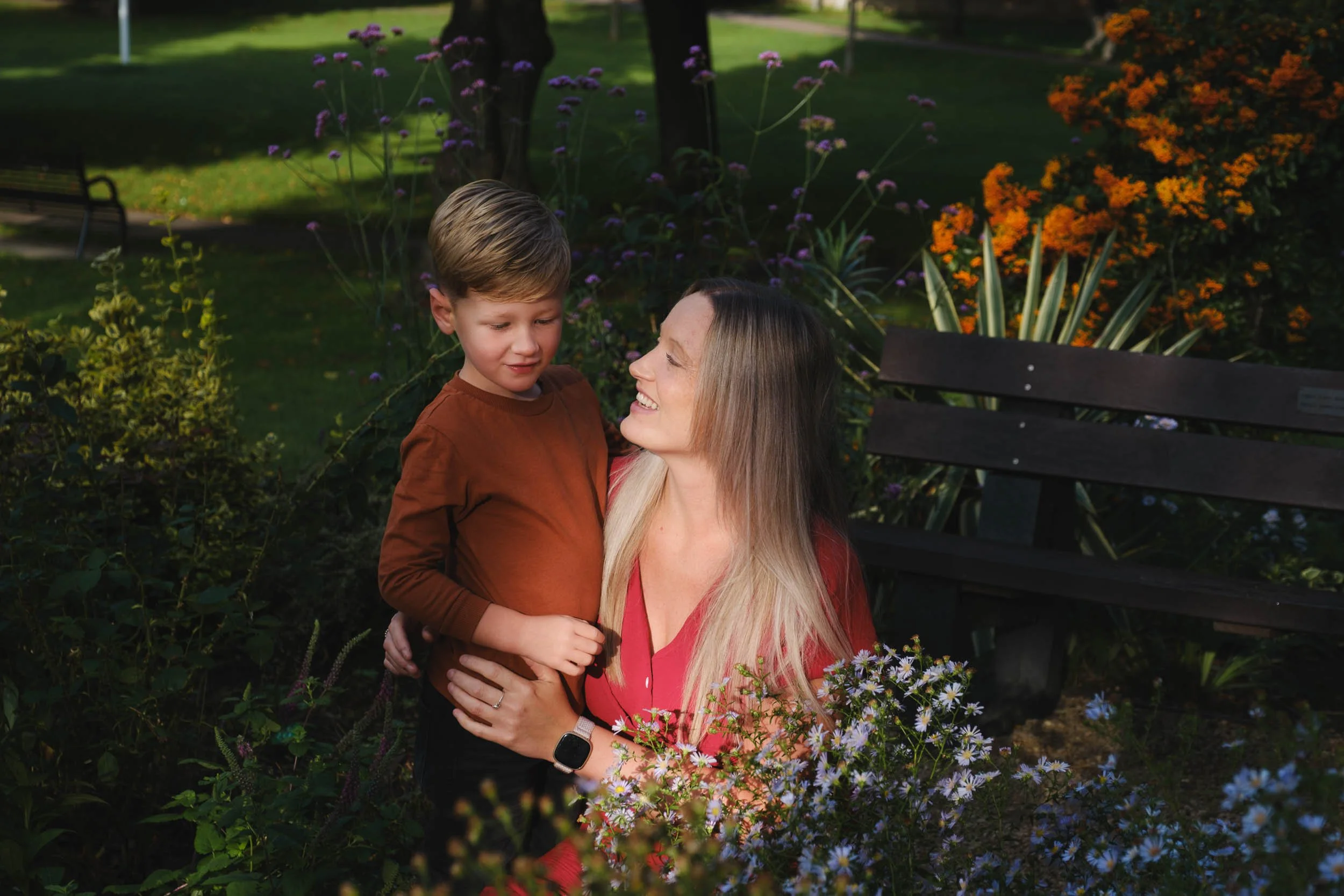 A woman holding a young boy in a garden with colorful flowers and green trees, smiling at each other.