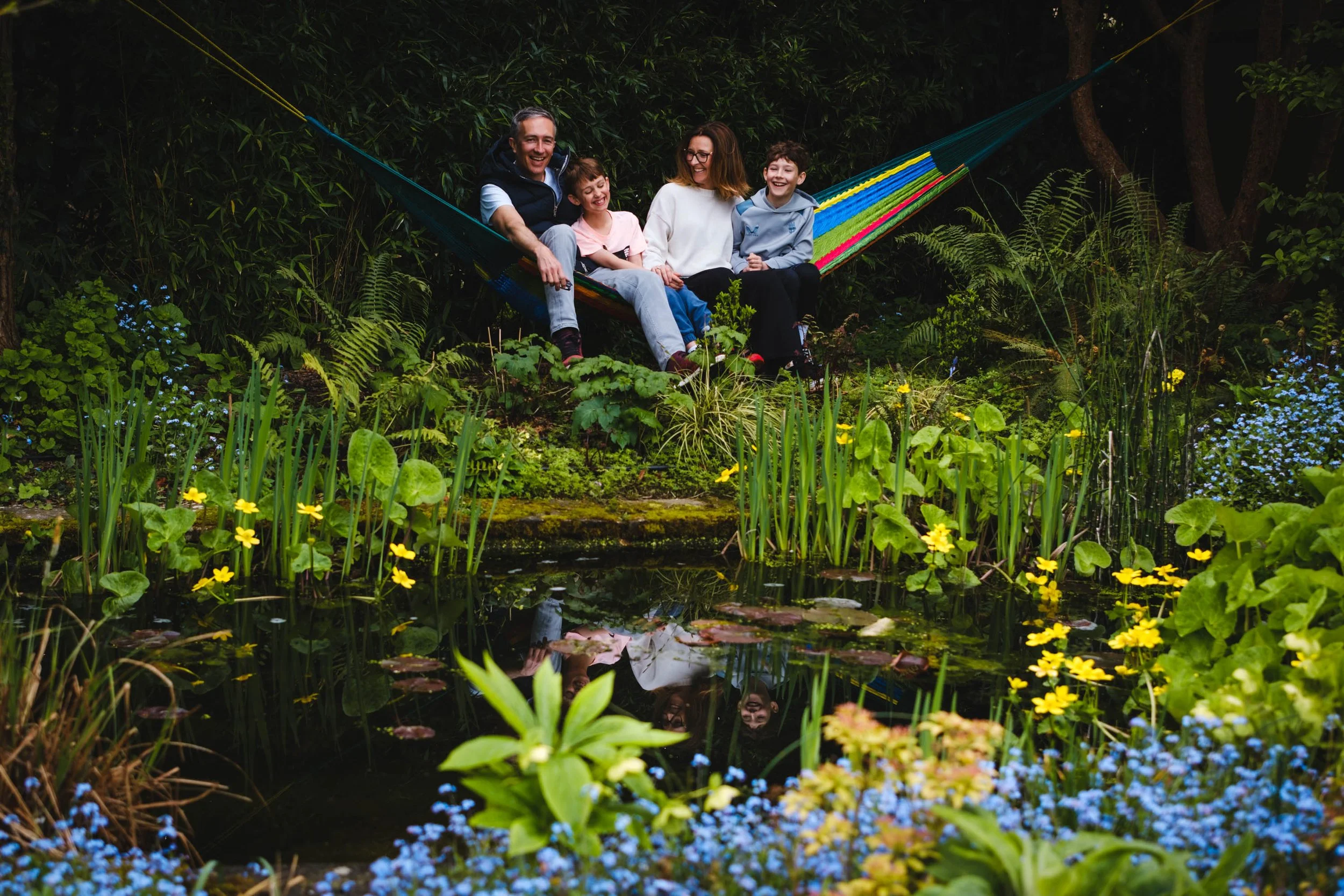 A family of five sitting on a colorful hammock in a lush, green garden, reflected in a pond with yellow and blue flowers.