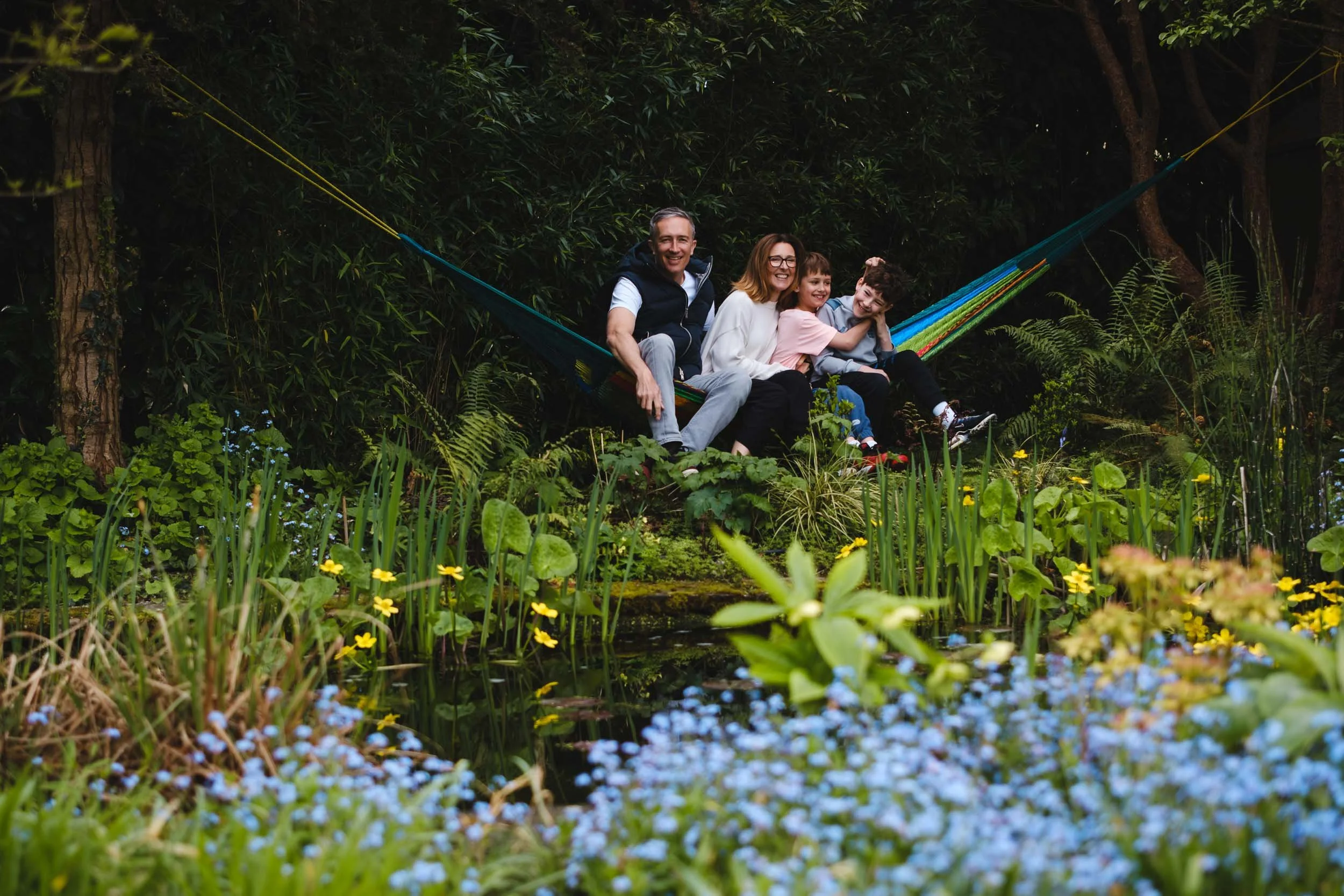 A family of four sitting on a hammock surrounded by lush greenery and blooming flowers in a garden.