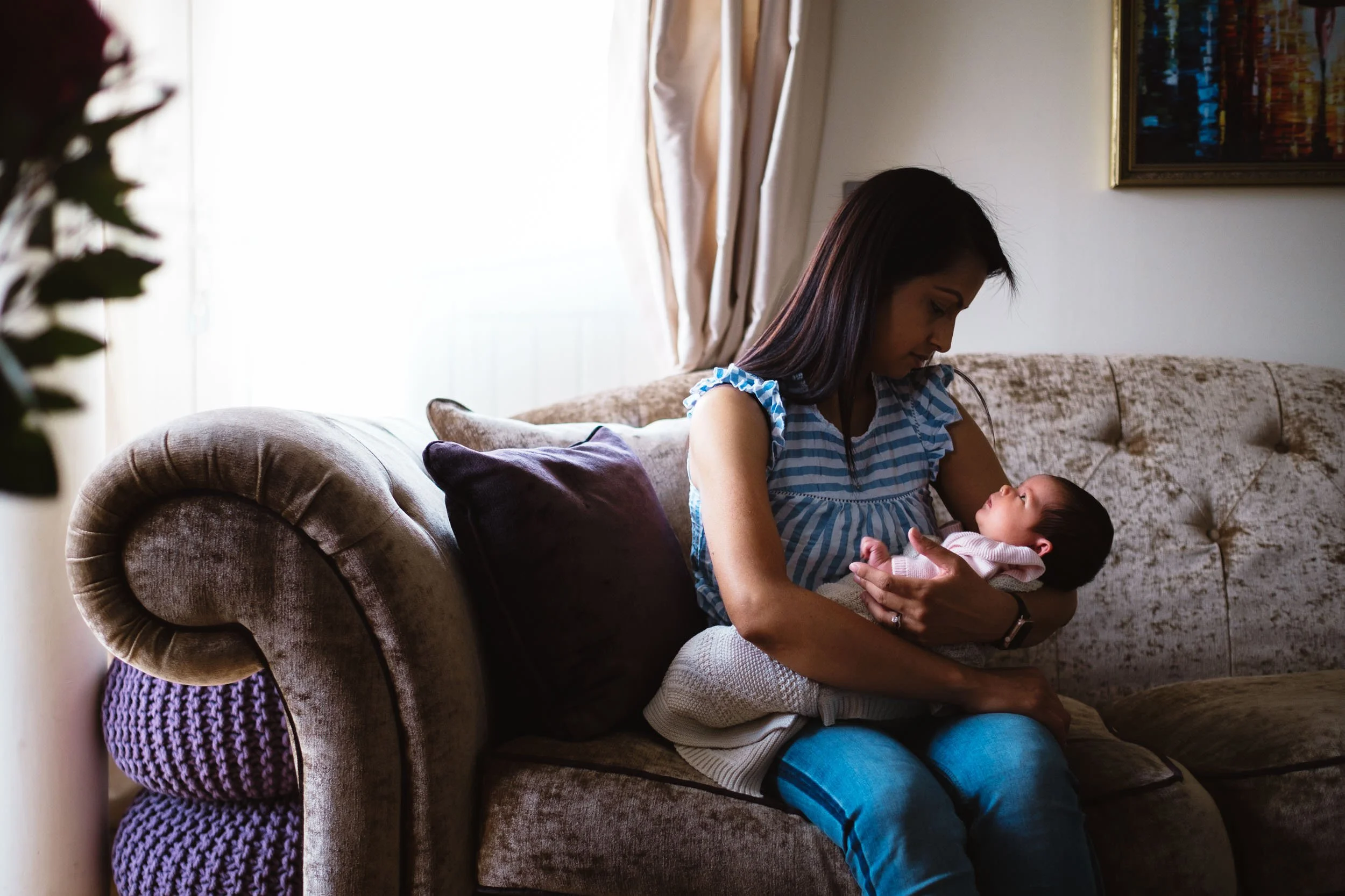 A woman sitting on a beige couch holding a baby in her arms inside a living room, with a window and curtains behind her and framed artwork on the wall.